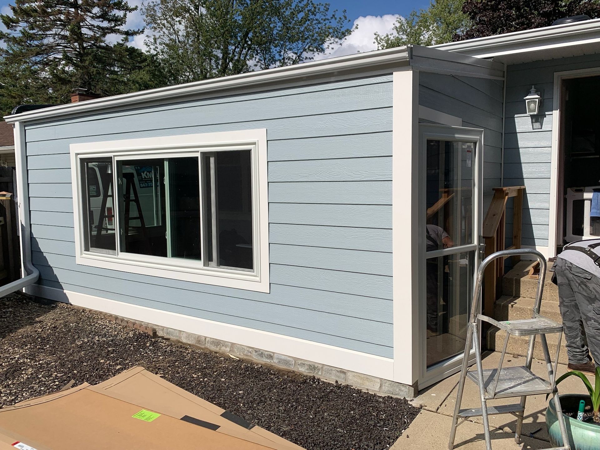 Light blue house addition with white trim, a large horizontal sliding window, a side door, and a metal ladder outdoors.
