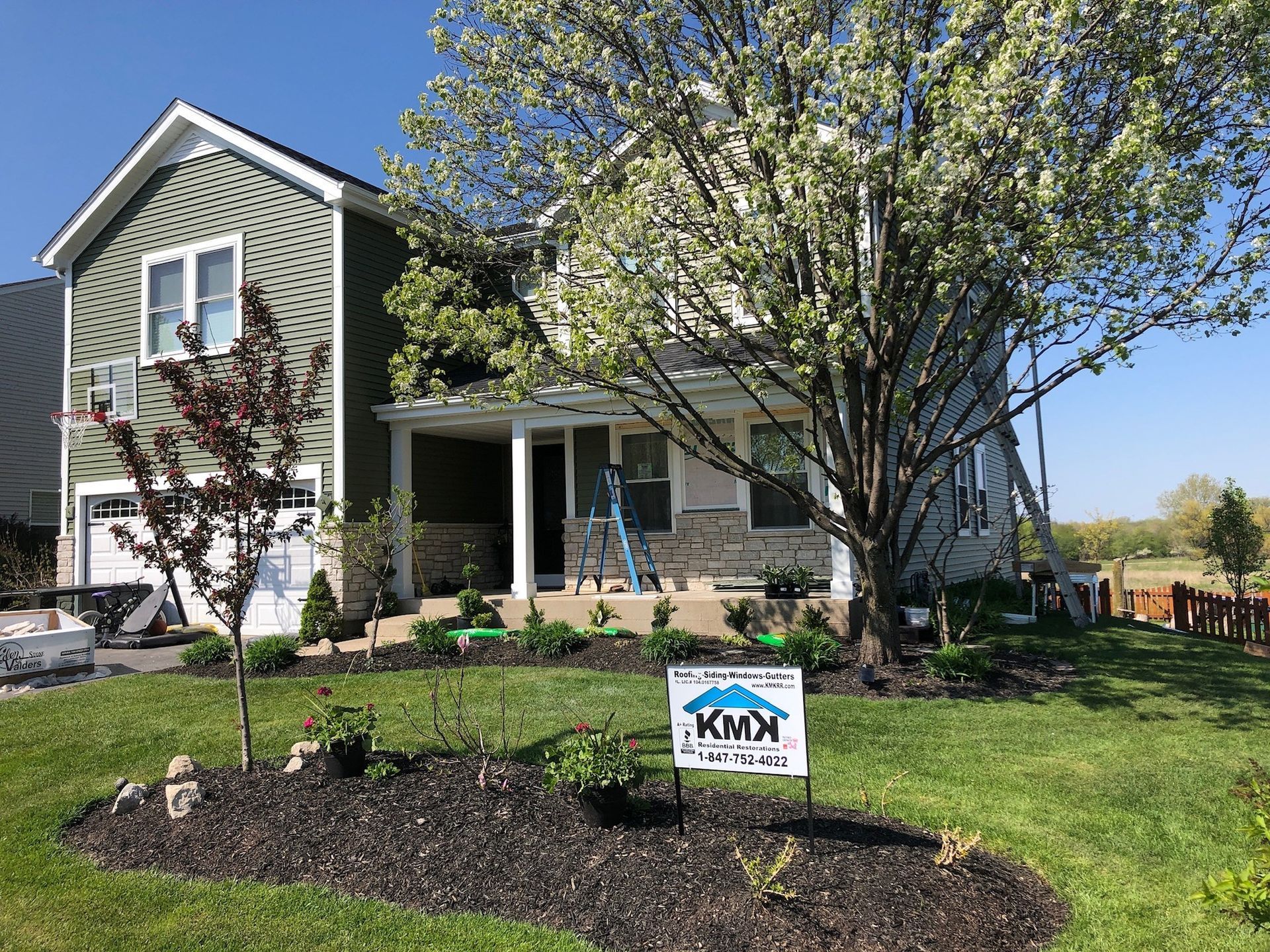 A two-story green house with a garage and a white porch, fronted by a landscaped yard with a blooming tree and a sign.
