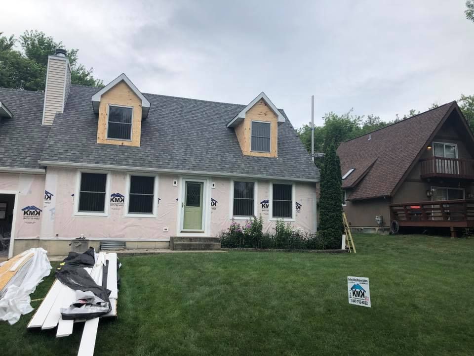 A light pink house under renovation with grey roof shingles, dormer windows, and construction materials on the lawn.