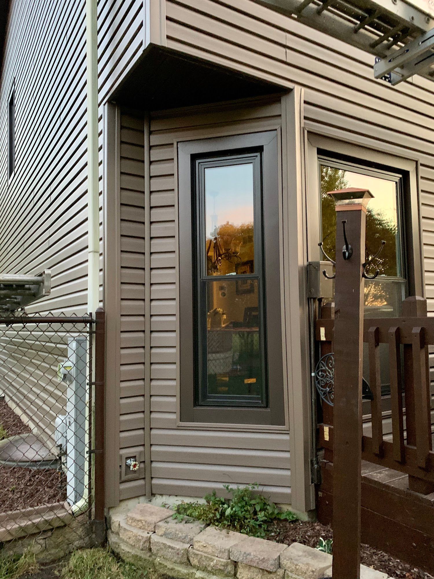 A narrow vertical window with a dark brown frame set into the corner of a house with horizontal brown siding.