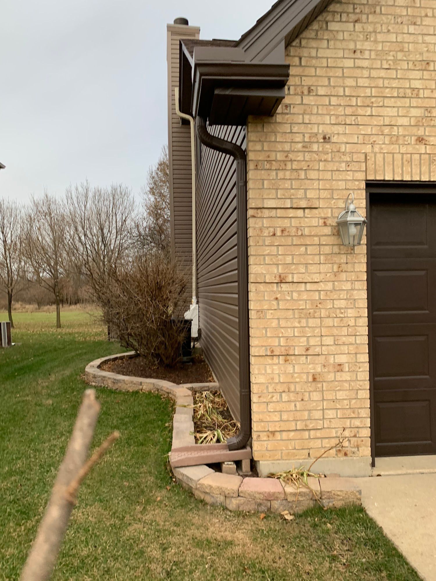 A side view of a brick house with brown siding, a dark downspout, and a landscaped stone flower bed near the lawn.