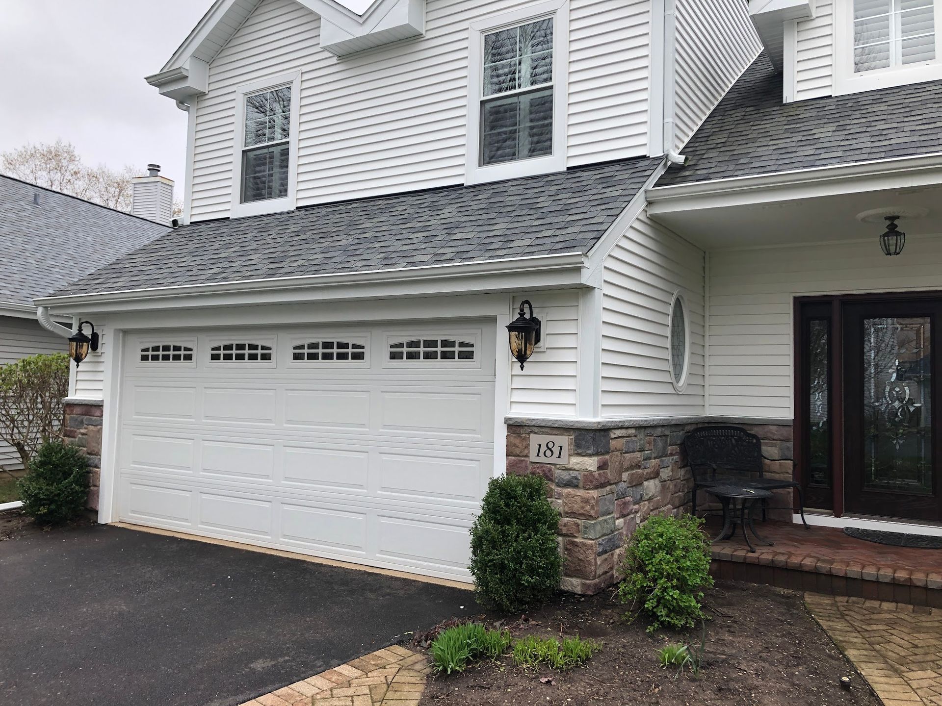 A white house with gray shingled roof, stone foundation, and a two-car garage with a front entryway on a cloudy day.