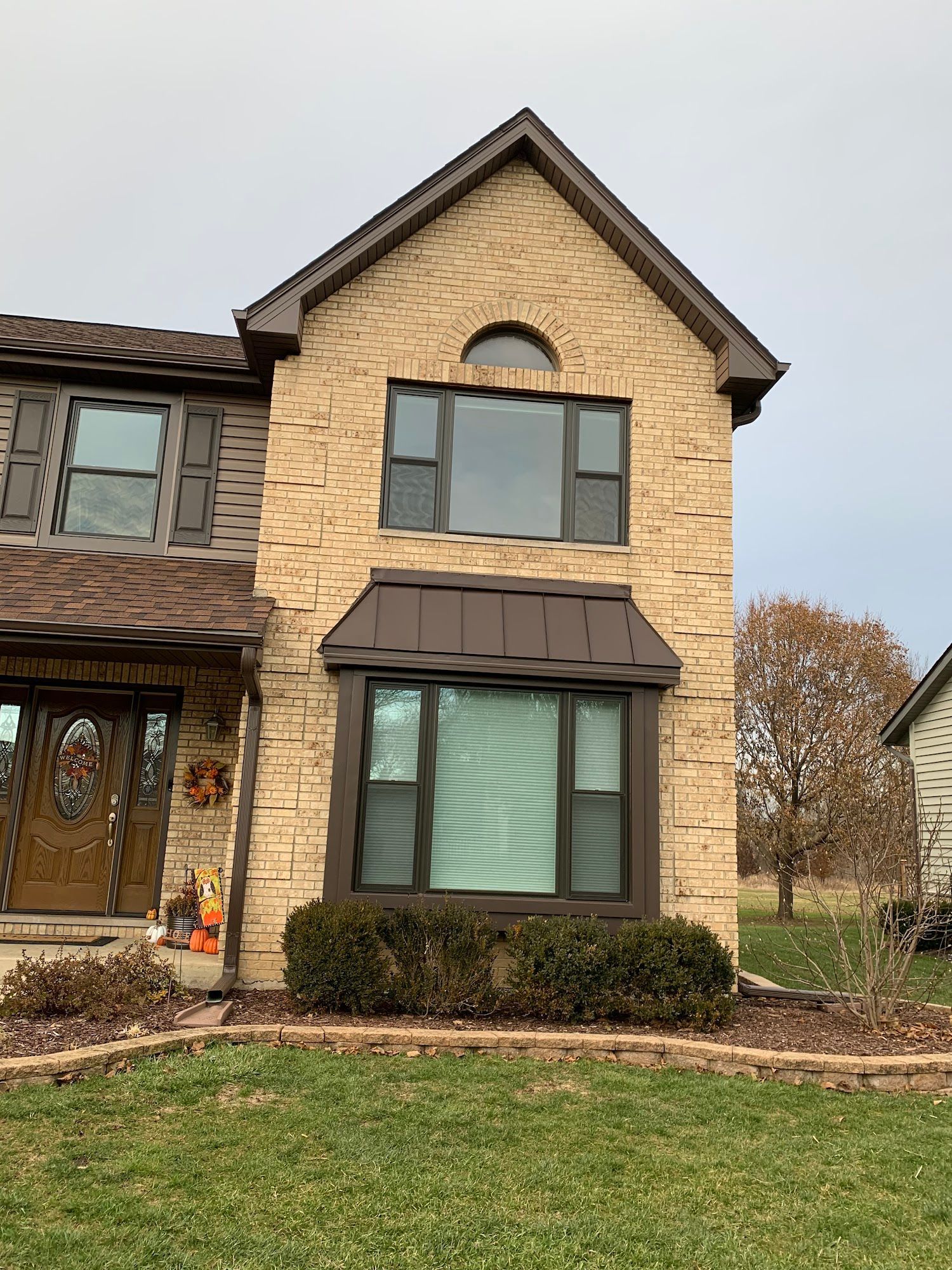 Tan stone facade of a two-story house with brown metal roofing, dark window frames, and a wooden front door.