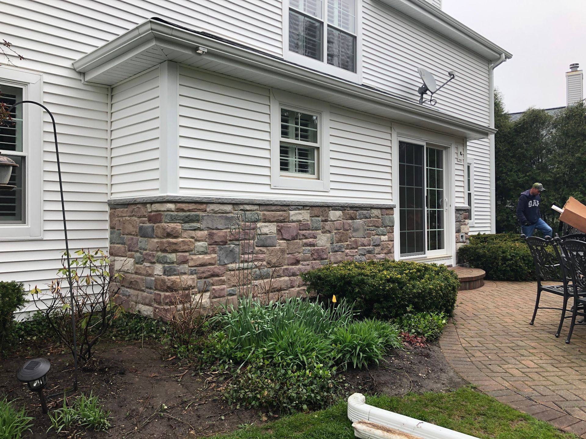 Exterior view of a house with white horizontal siding and a stone-veneer base, featuring a patio door and landscaping.