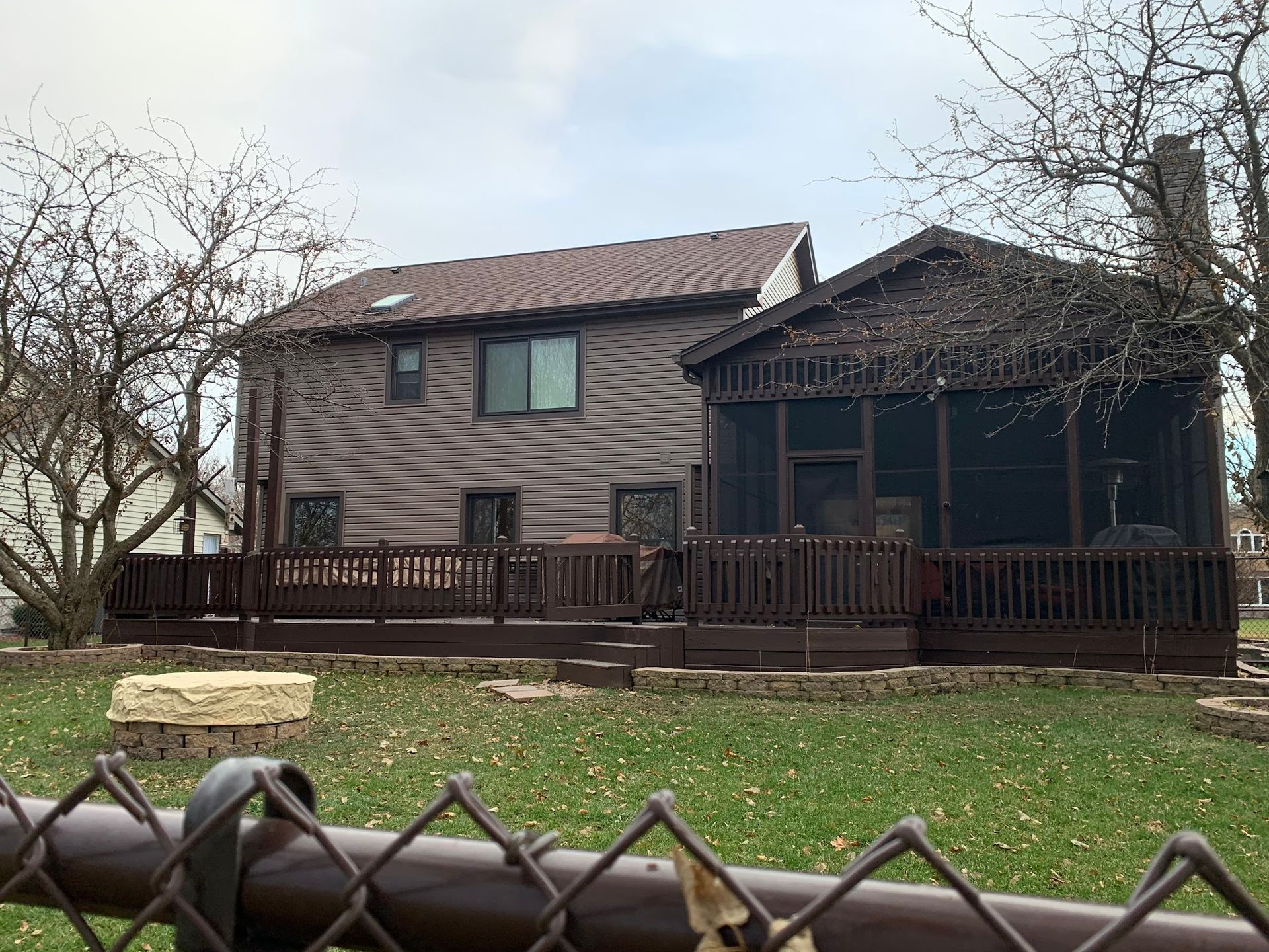 Two-story house with brown siding and a large, elevated deck featuring a screened-in porch, set in a grassy backyard.