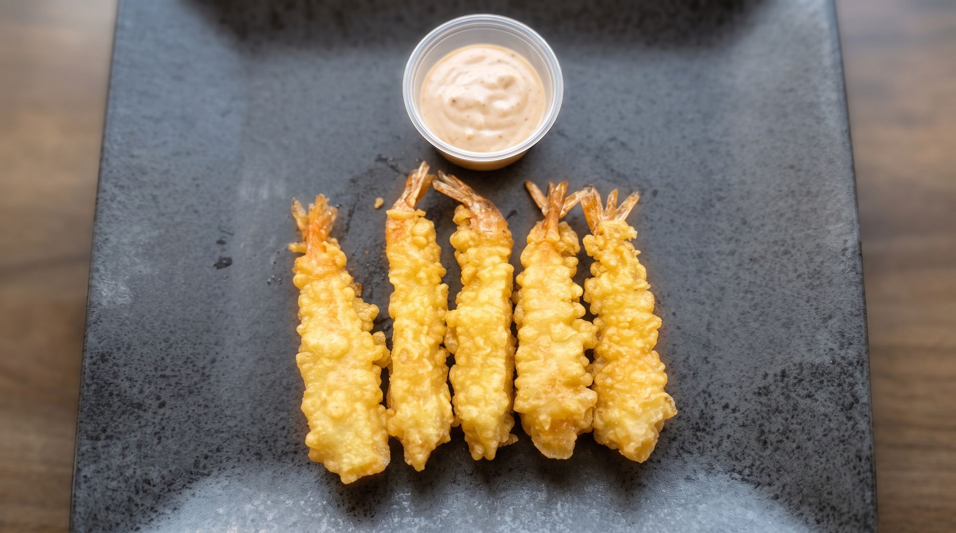 Four golden fried shrimp on a black plate with a small cup of dipping sauce