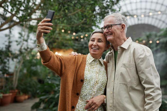 Elderly couple taking a selfie outdoors in a lush green garden, both smiling with arm around each other.