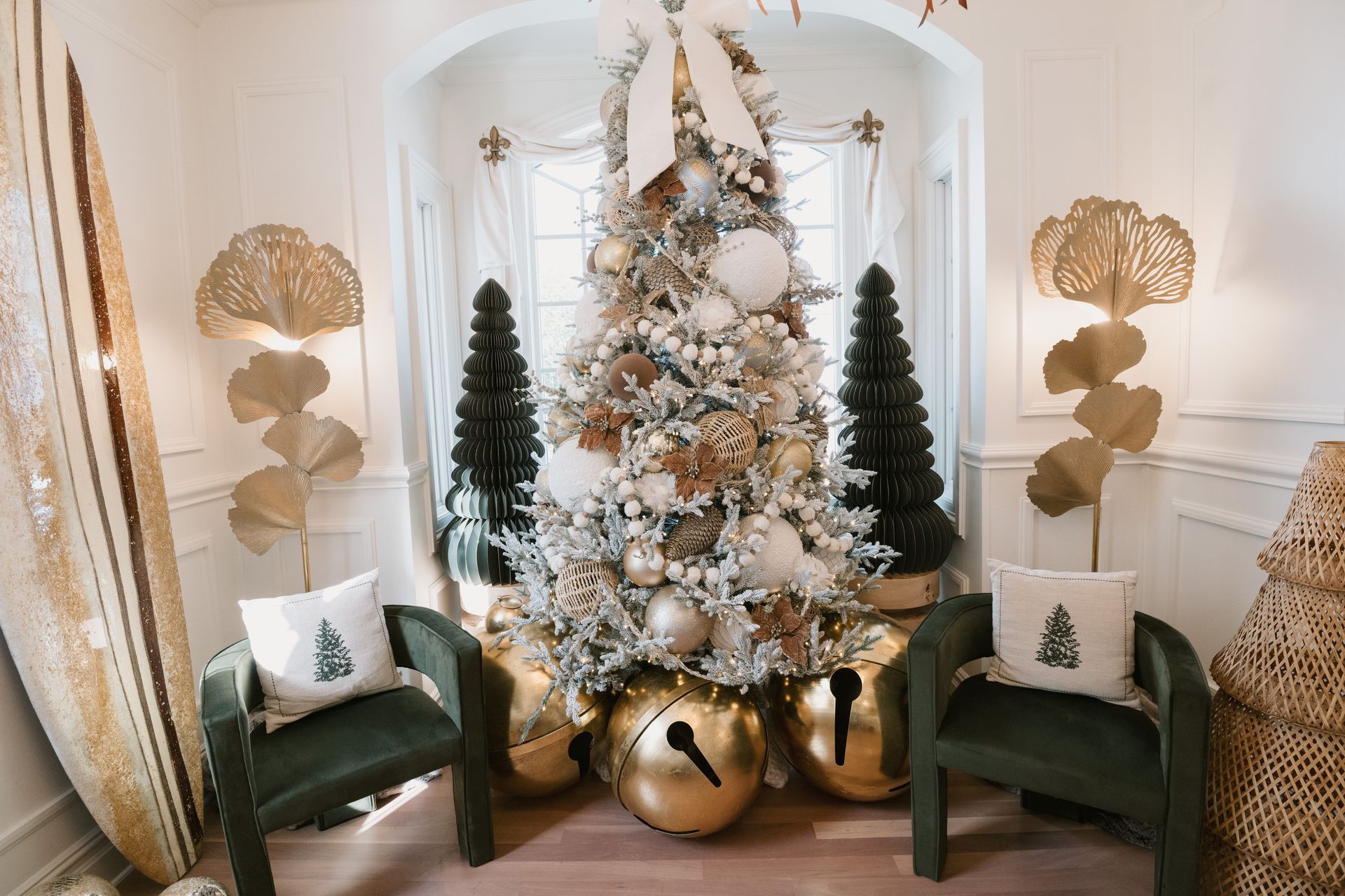 Christmas tree display with gold accents, green chairs, and snowflake pillows in a bright room.