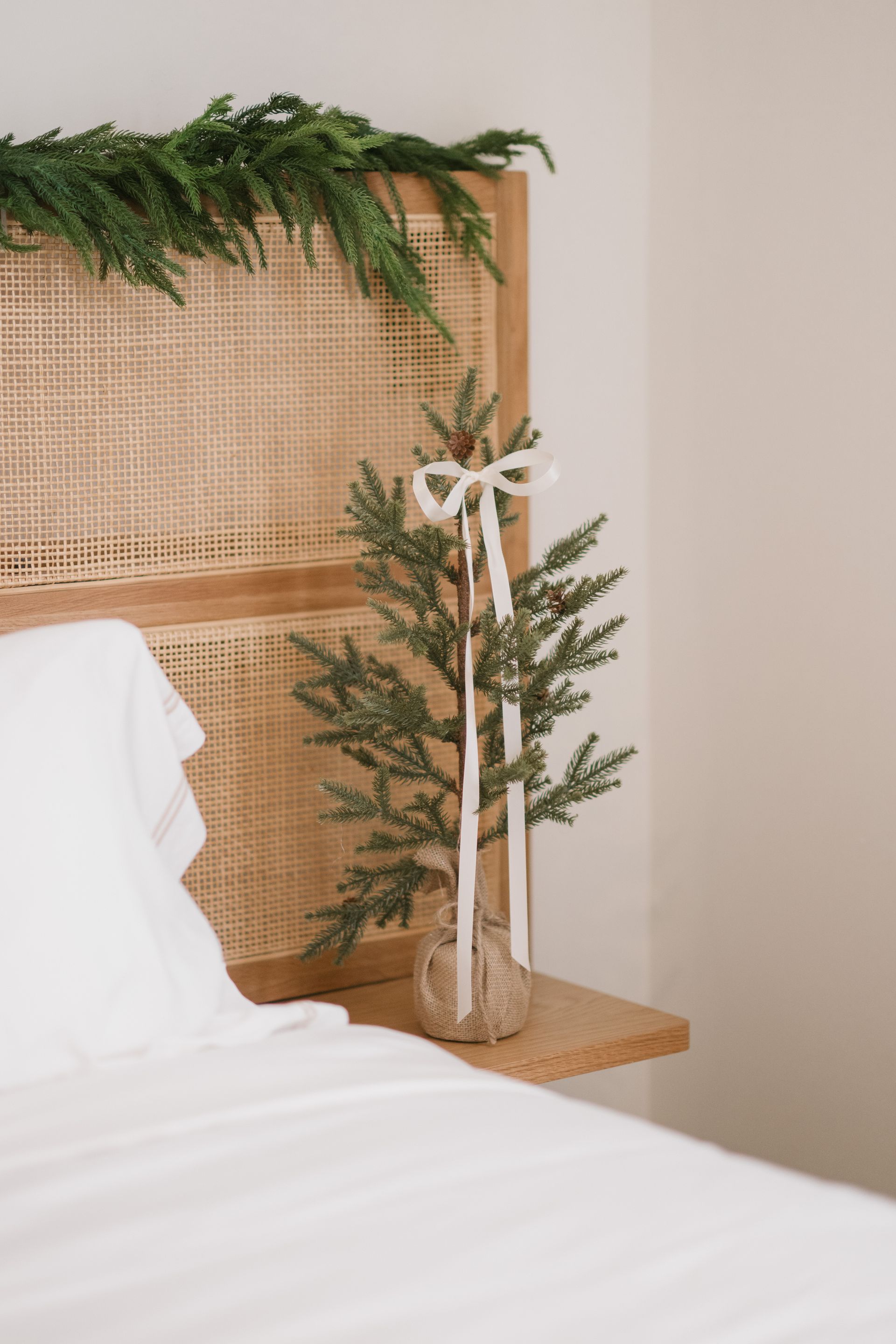 Christmas decor: Small tree with ribbon on a wood nightstand, bed with white linens, green garland on headboard.