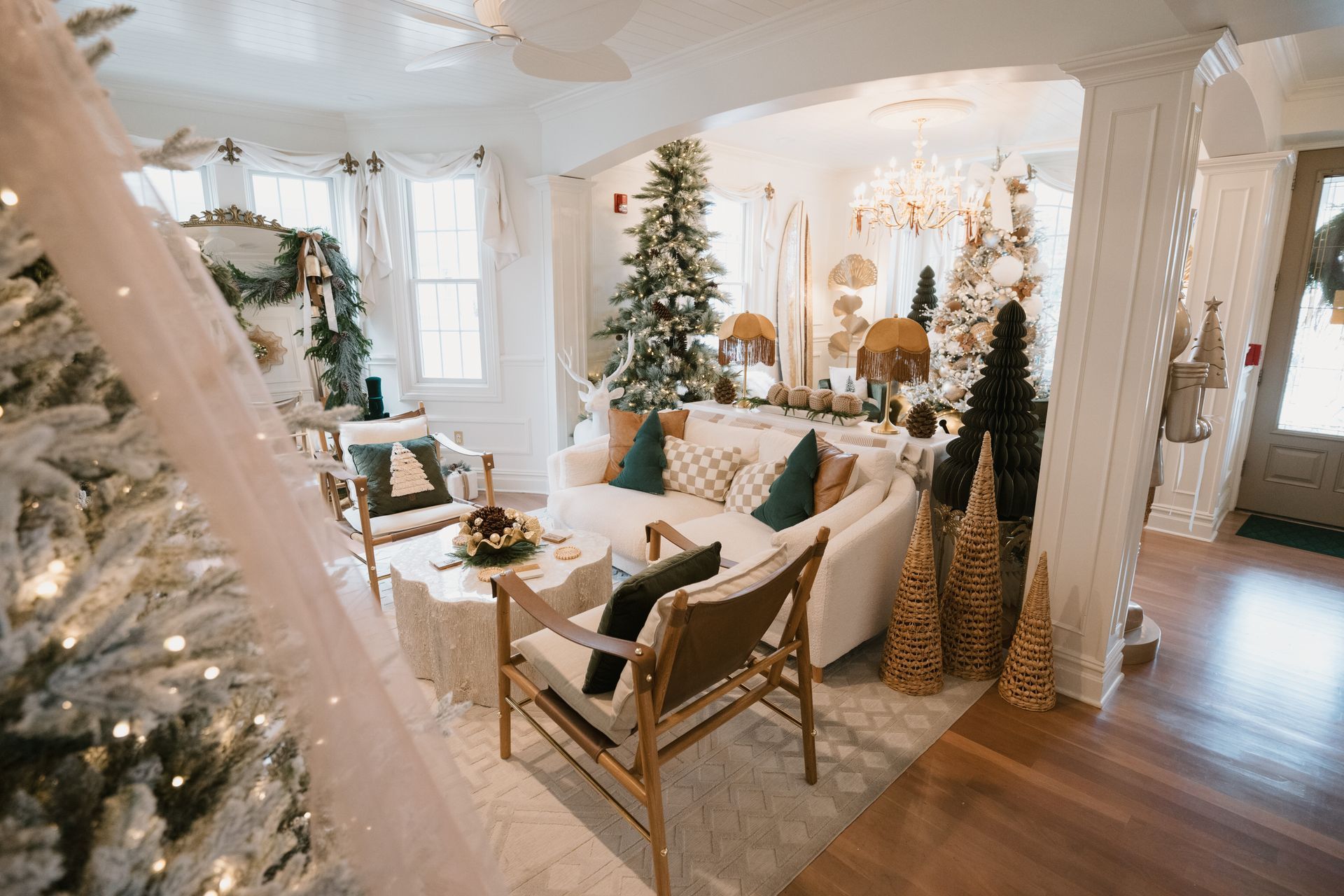 Living room decorated for Christmas with multiple trees, white sofas, and festive decor.