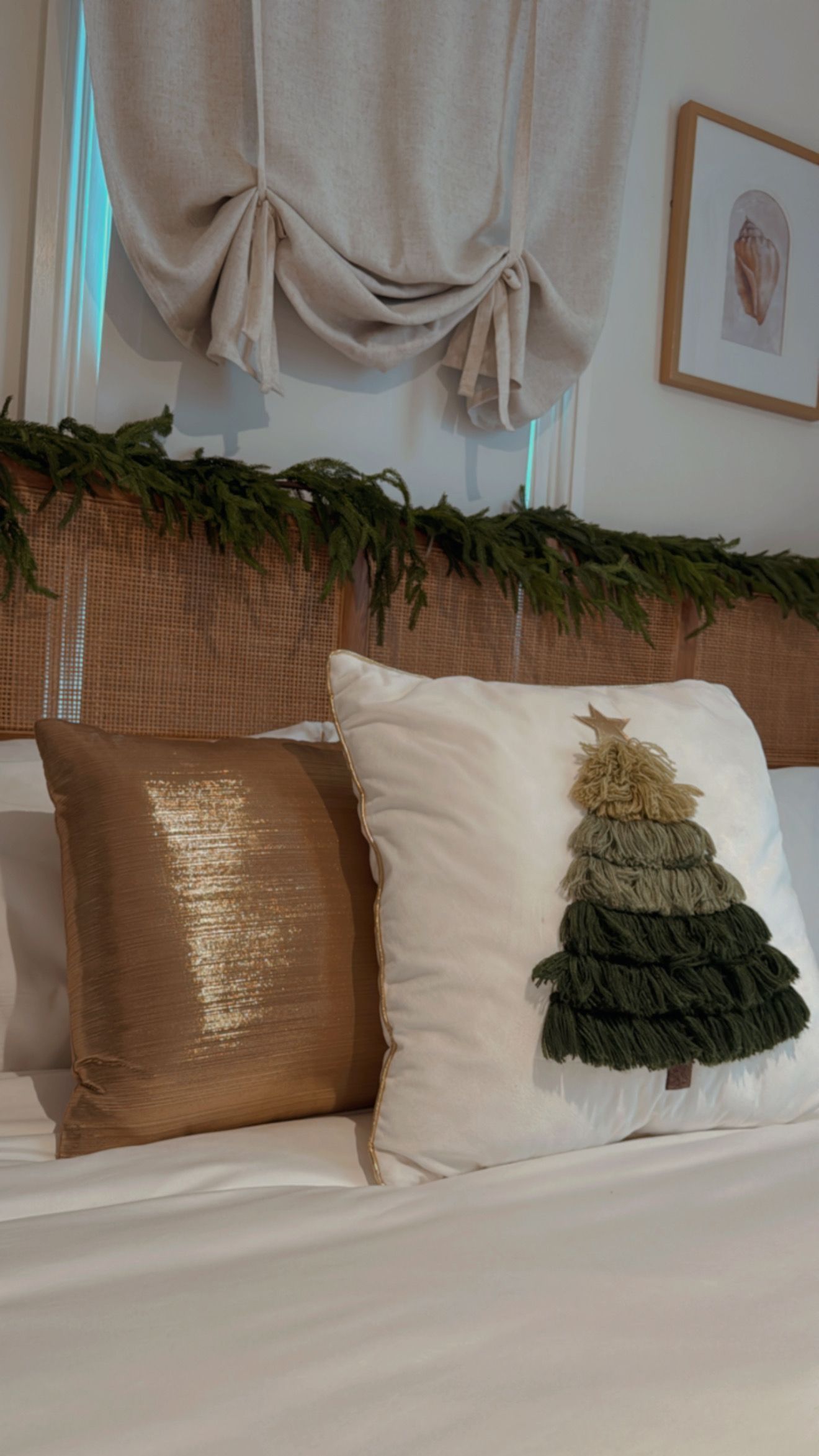 Bed with gold sequin and Christmas tree pillows, green garland, beige curtains, and framed art.