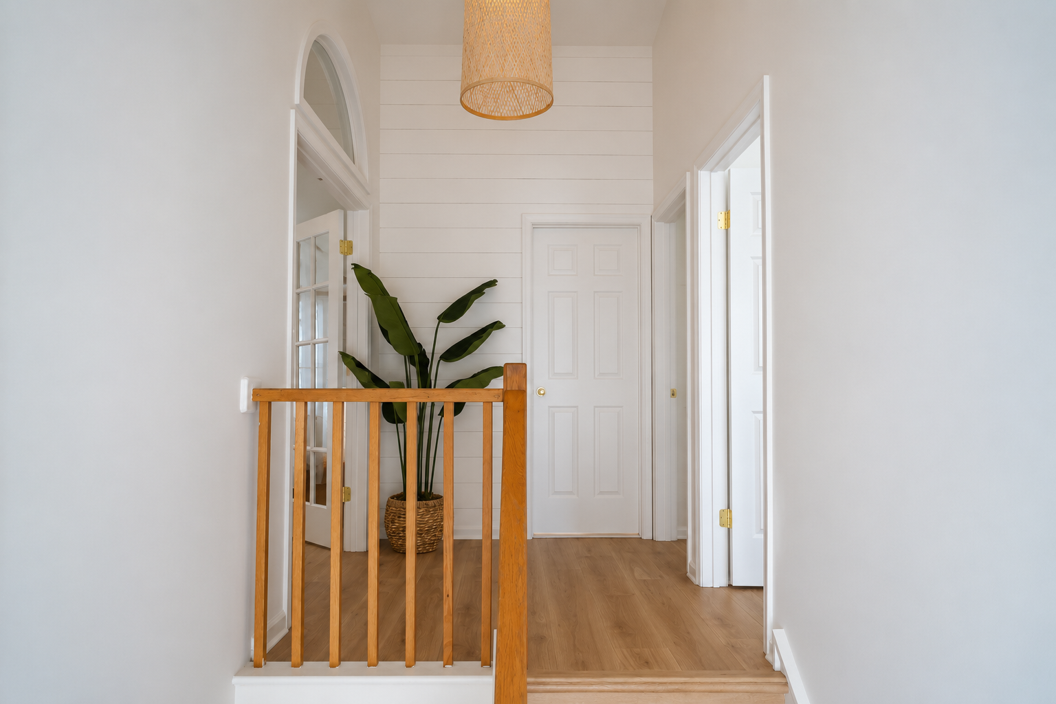 Bright hallway with white walls, wood floor, a potted plant, and a wooden stair railing.