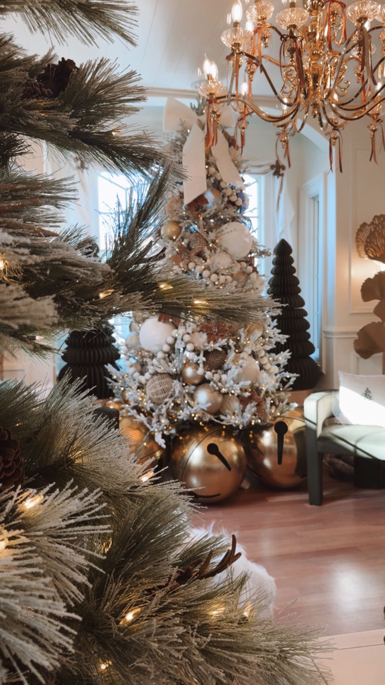 Christmas tree decorated with gold, white, and silver ornaments, with golden bells and a chandelier in the background.