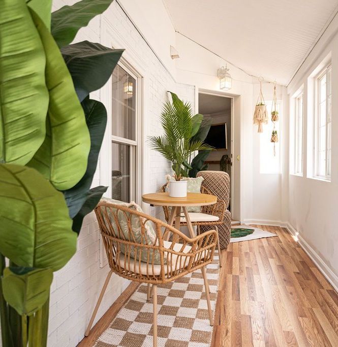 Cozy sunroom with rattan furniture, potted plants, and checkered rug. White walls, wood floor.