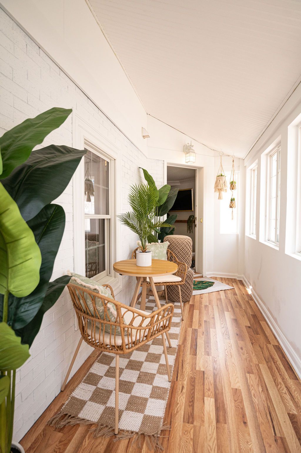 Sunroom with wood floors, white walls, plants, wicker furniture, and a checkered rug.