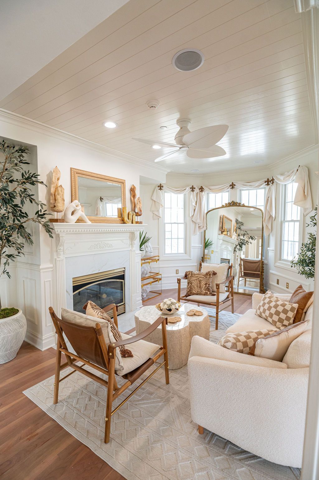Cozy, bright living room with white fireplace, cream sofa, wood chairs, light rug, and large windows.