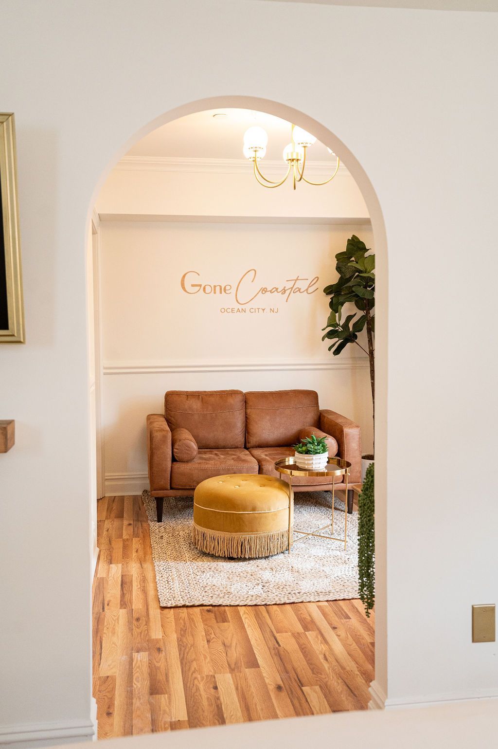 Interior archway frames a room with a brown couch, rug, and wooden floor. A round ottoman sits in front of the couch.