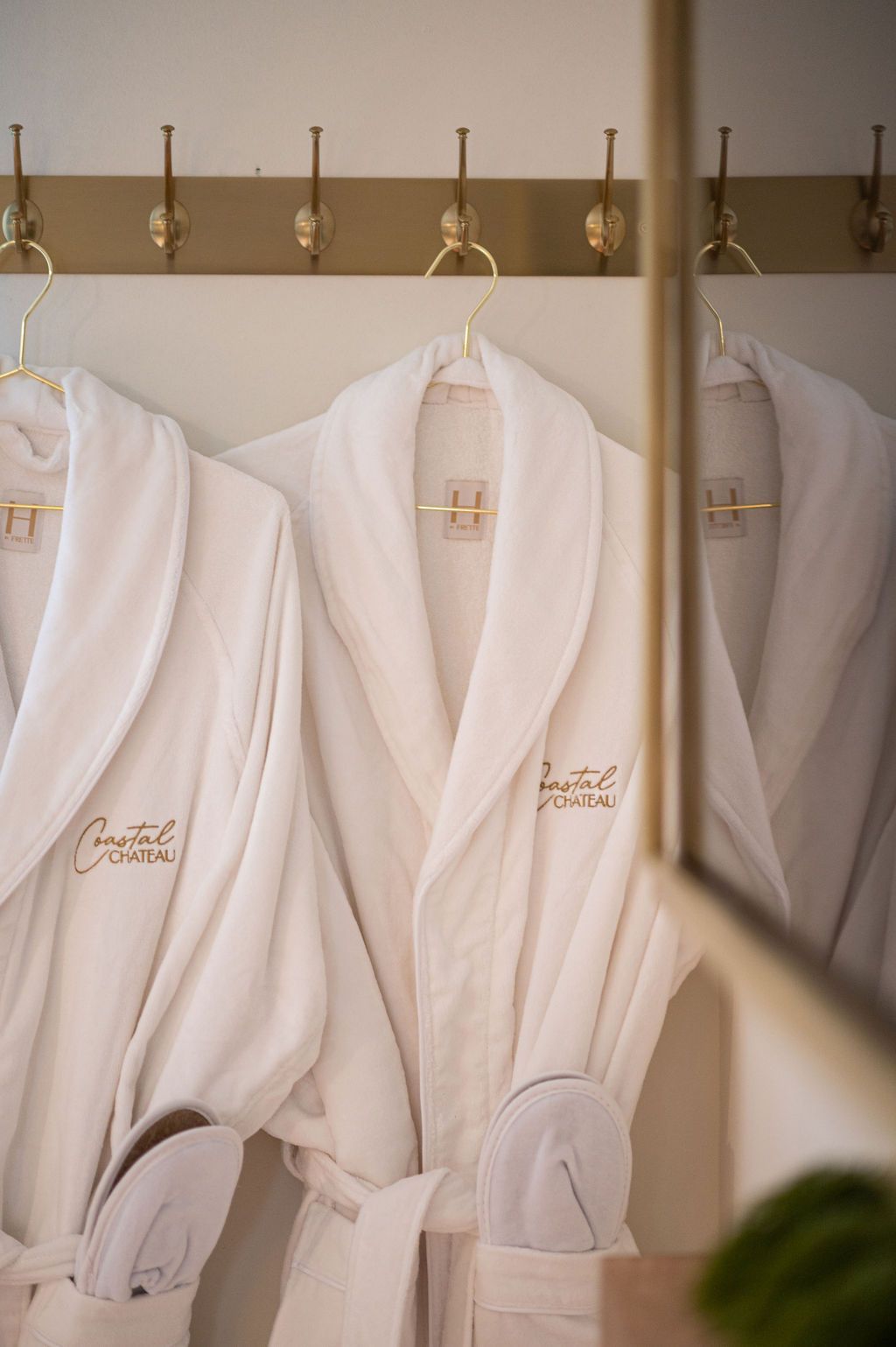 White bathrobes hanging on hooks with gold lettering and a mirrored reflection.