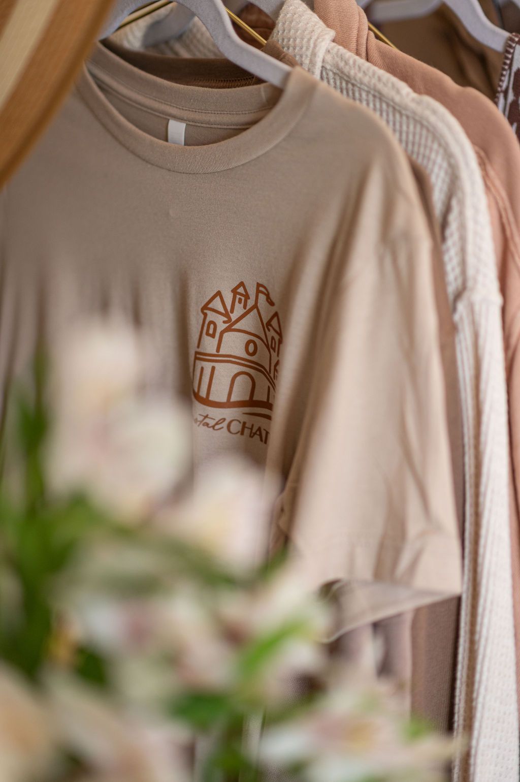 Beige t-shirt with brown castle graphic hangs among other neutral-colored clothes in a closet.