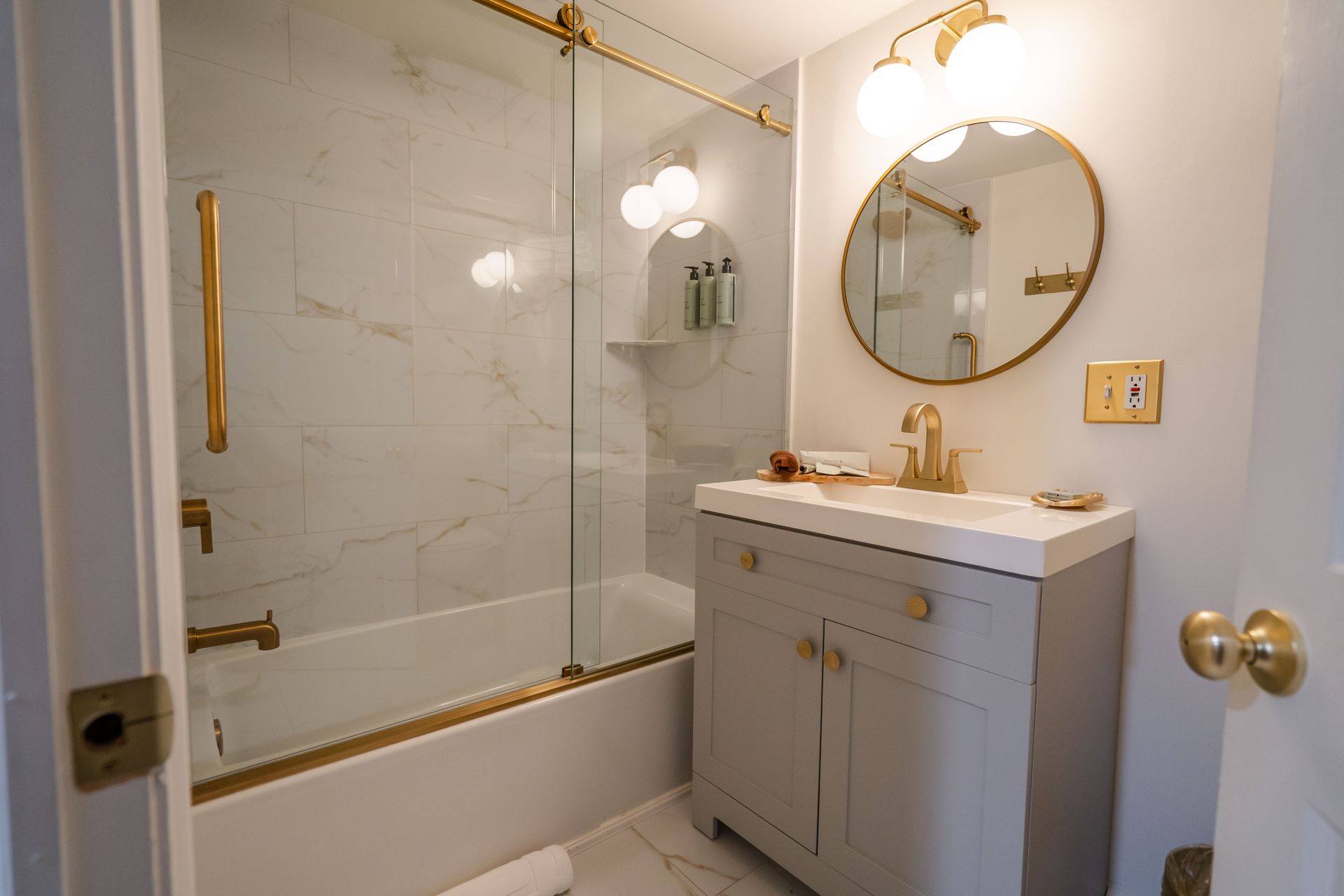 Bathroom with gold fixtures, marble-look tile, glass shower door, light gray vanity, and round mirror.