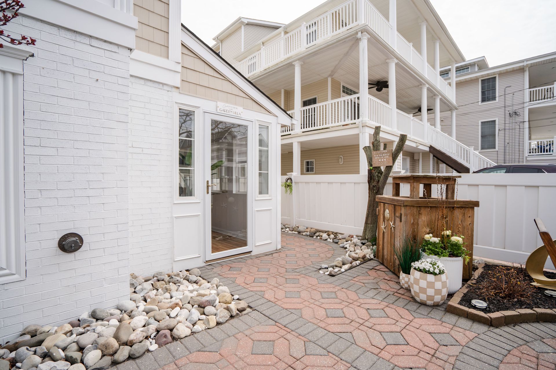 Backyard patio with brick paving, white and gray rocks, and a building with white siding.