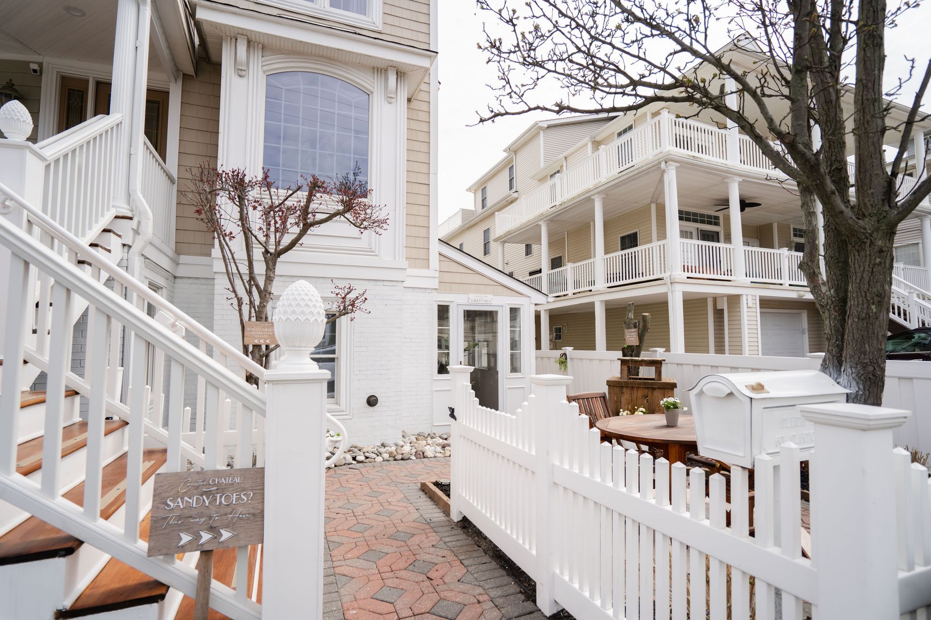 White picket fence and stairs lead to a light-colored Victorian house with a balcony and a decorative topiary.