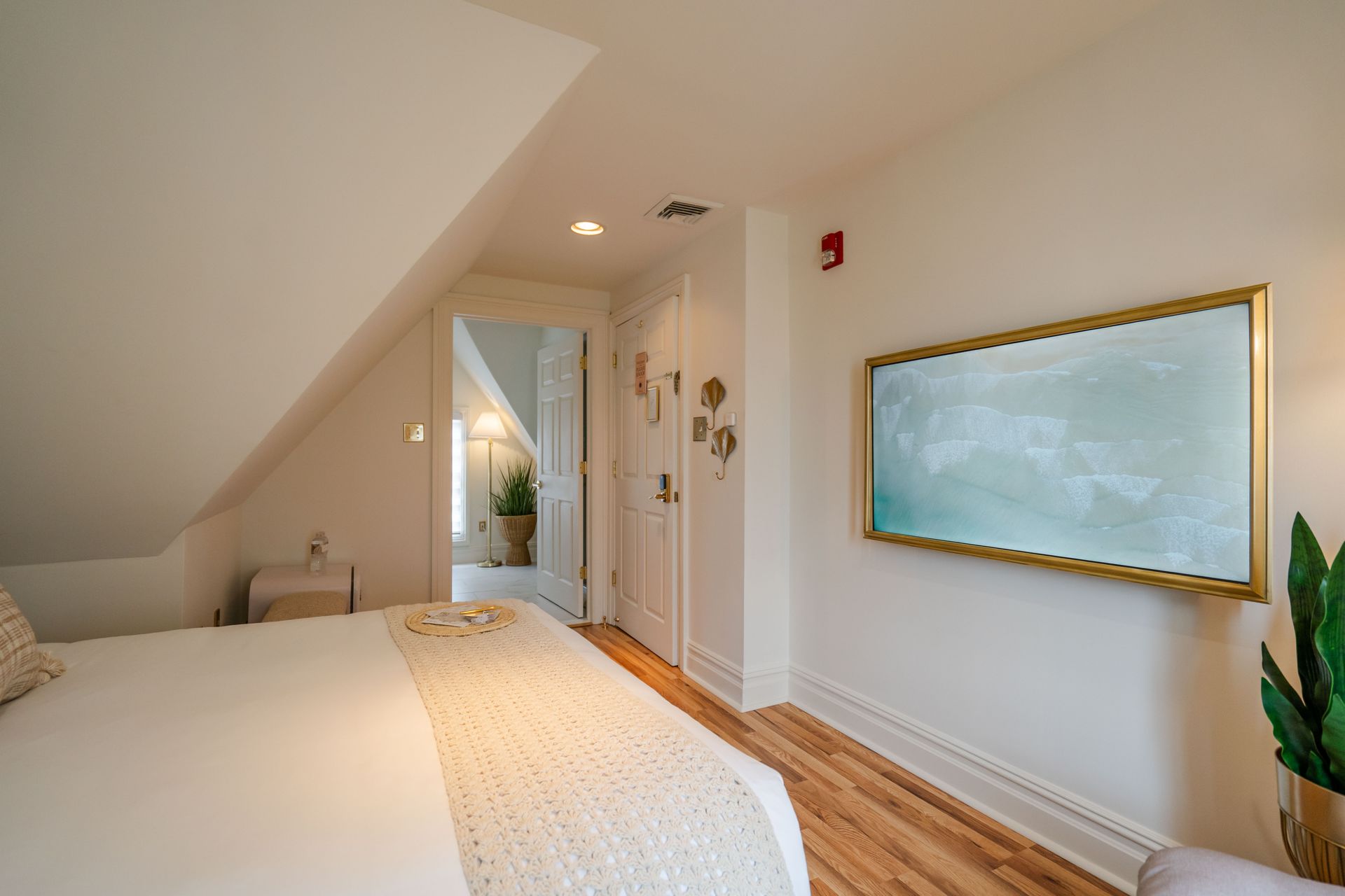Bedroom with white walls, angled ceiling, bed, art, door to another room.