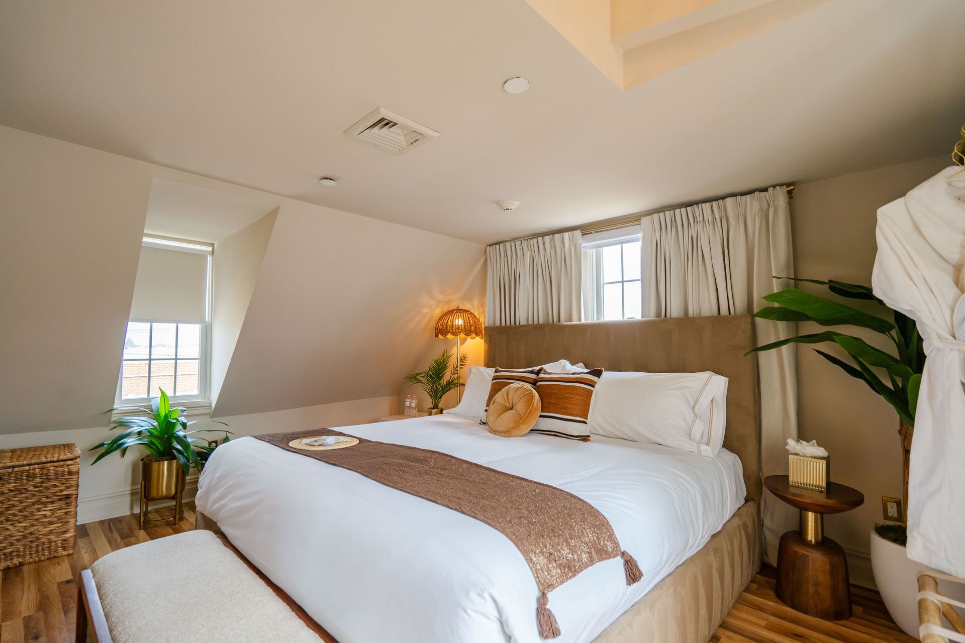 Bedroom with a bed, bench, plants, and small windows with white curtains.