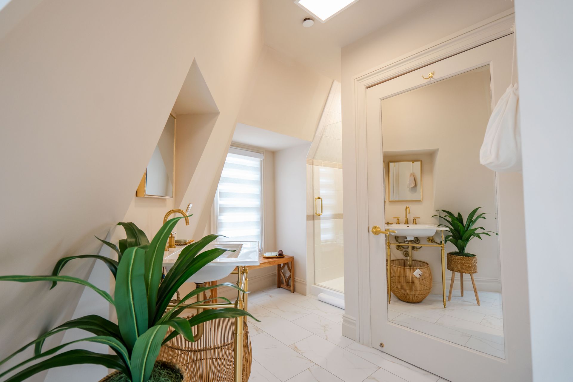 Elegant, light-filled bathroom with gold fixtures and plants. Features a large mirror and a shower stall.