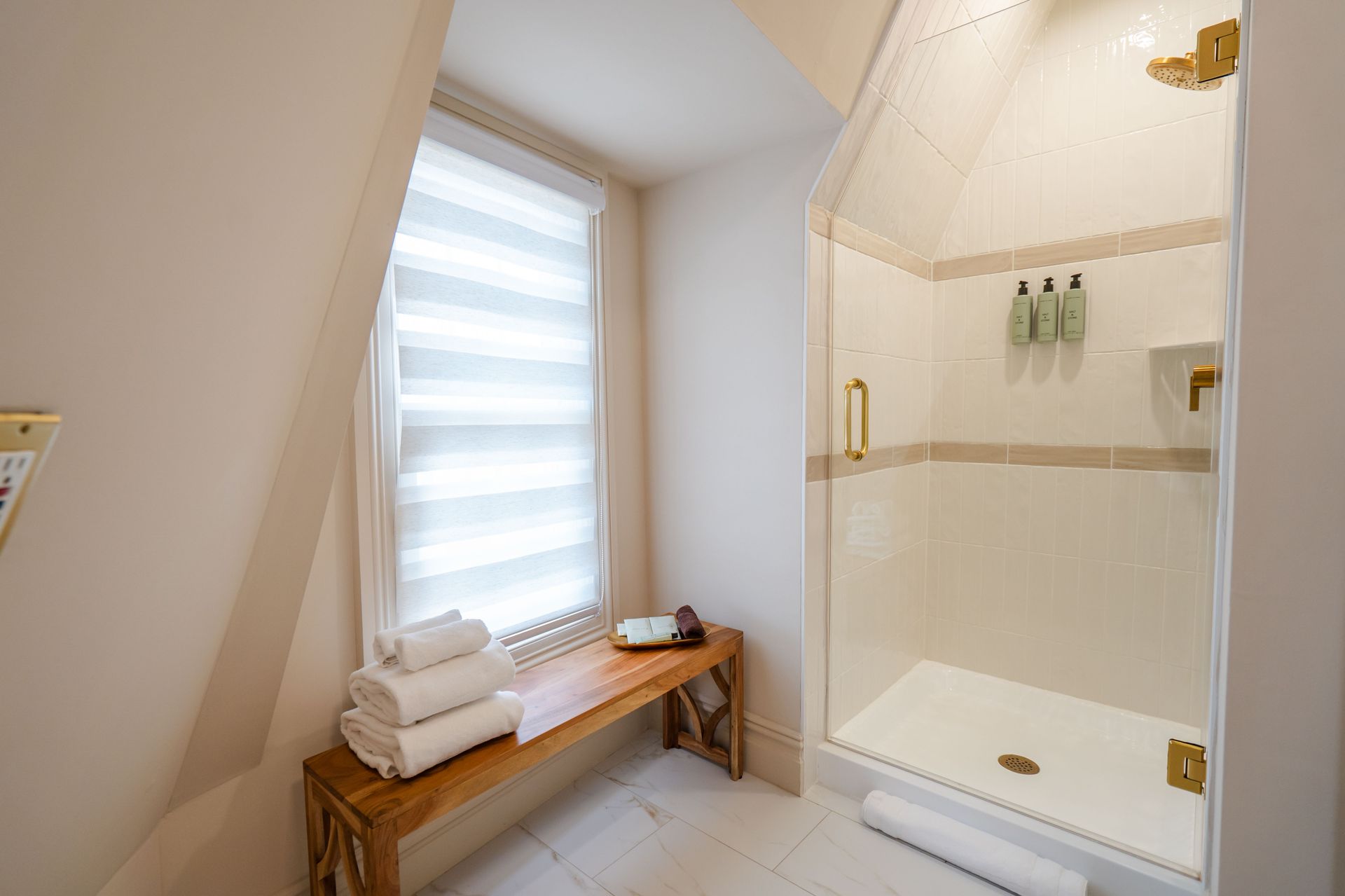 Bathroom with angled ceiling, shower, window with blinds, towels, and a wooden bench.