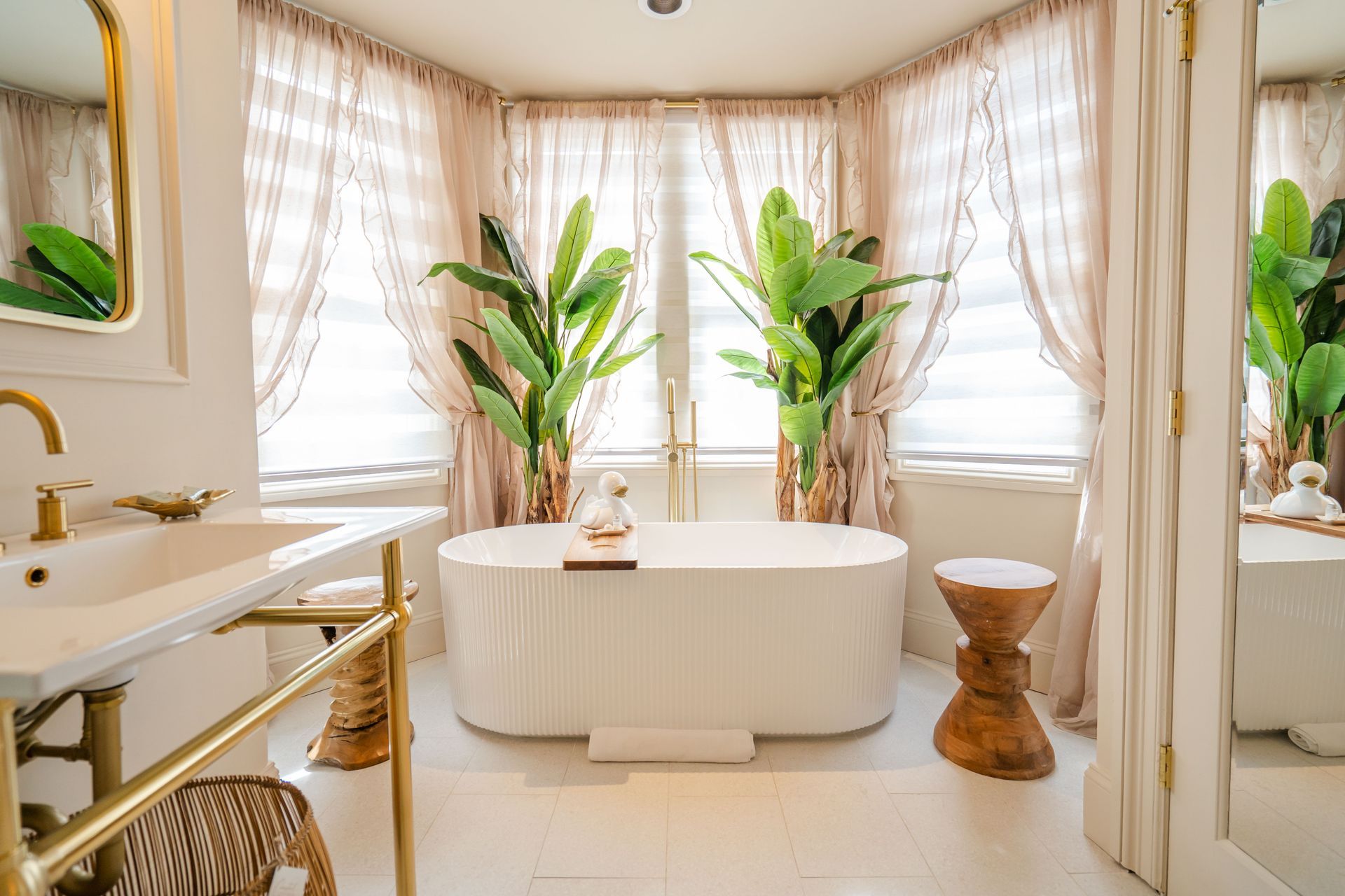 Elegant white bathroom with a freestanding tub, gold accents, and plants by a window.