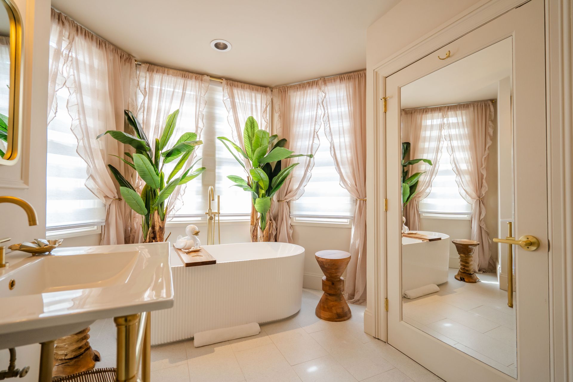 Elegant bathroom with a free-standing tub, large windows with sheer curtains, and a gold-accented sink.