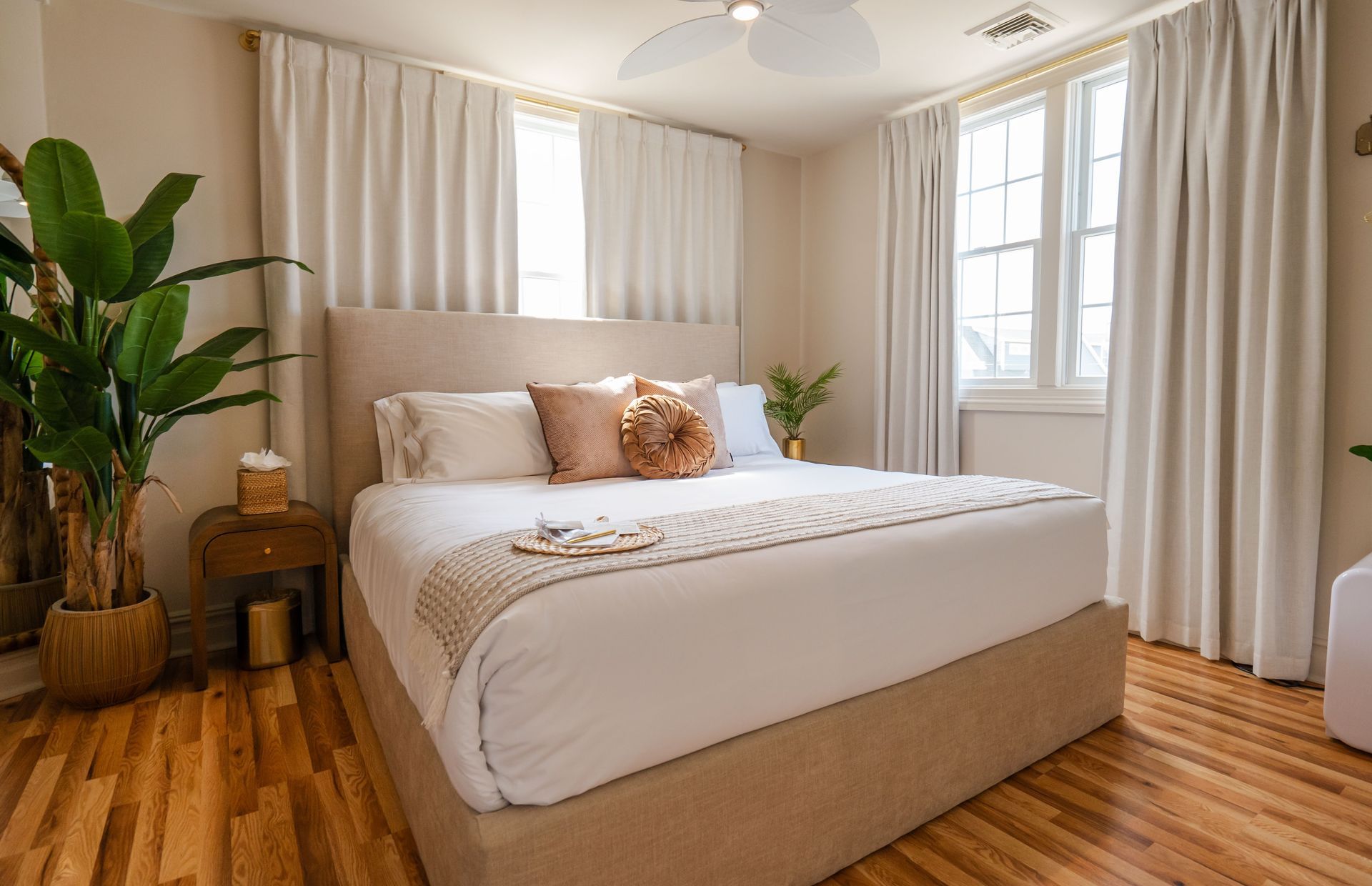 Bedroom with a bed, plants, light-colored curtains, and hardwood floors.