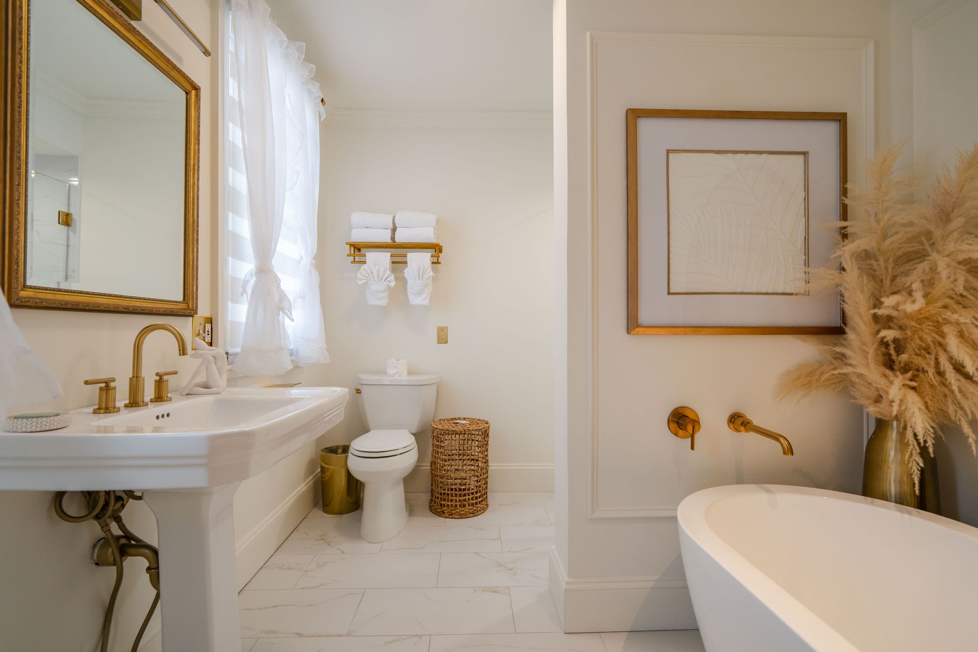 Bright bathroom with white and gold accents, including a sink, toilet, bathtub, and decorative pampas grass.