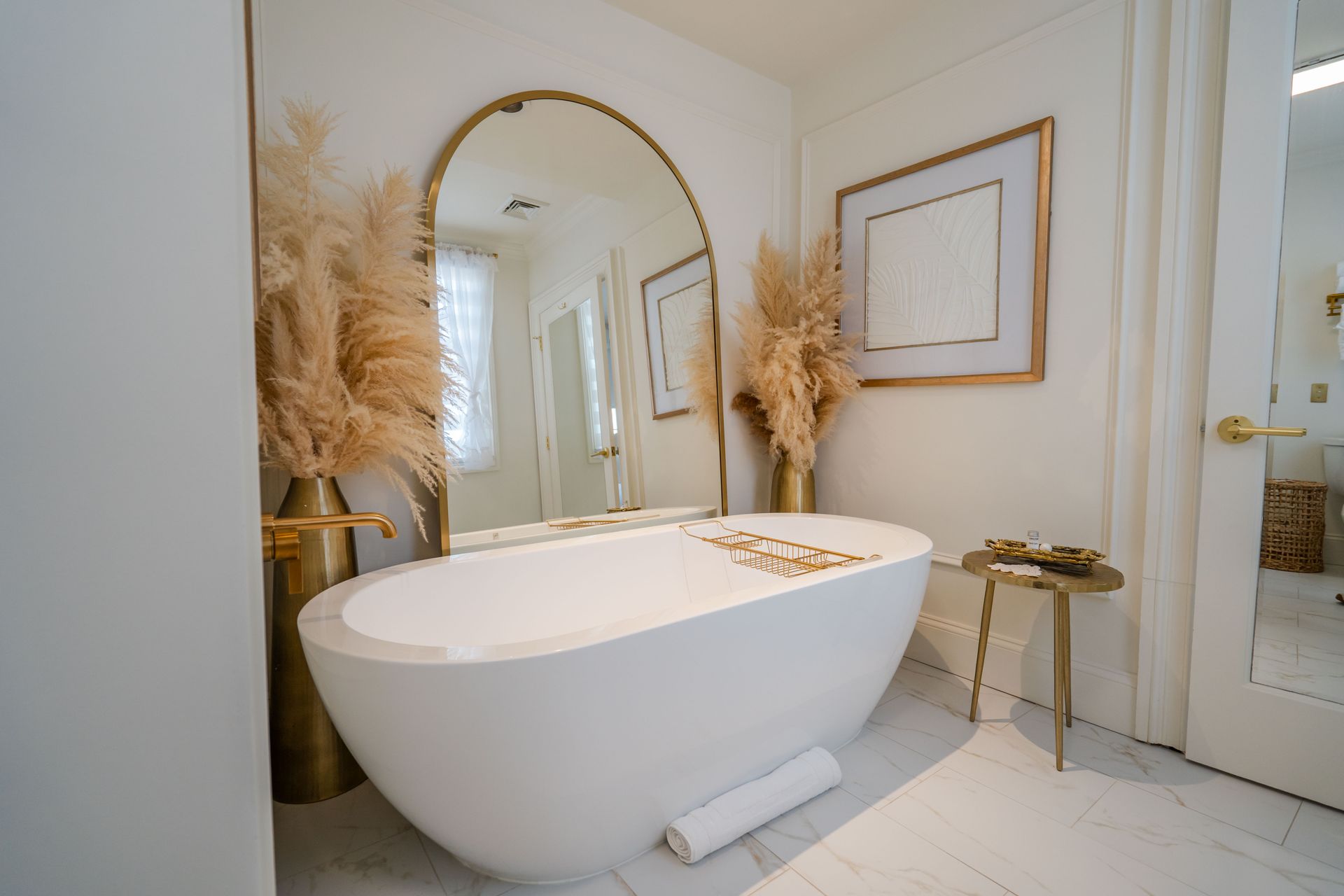 White bathtub in a modern bathroom with gold accents, mirror, pampas grass, and side table.