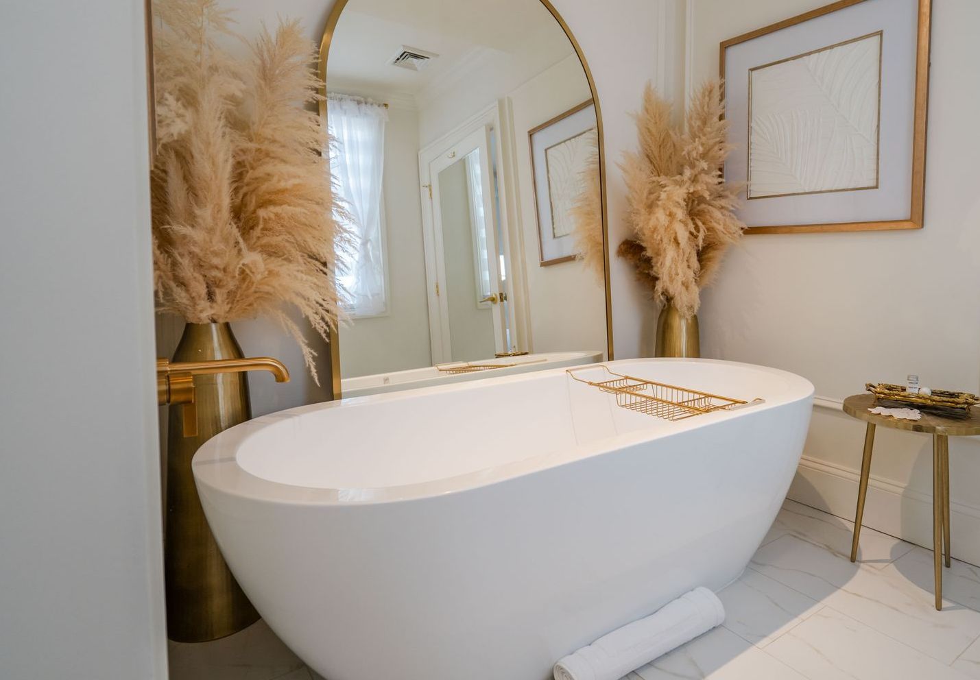 White, modern bathroom with freestanding tub, gold accents, pampas grass, and large mirror.