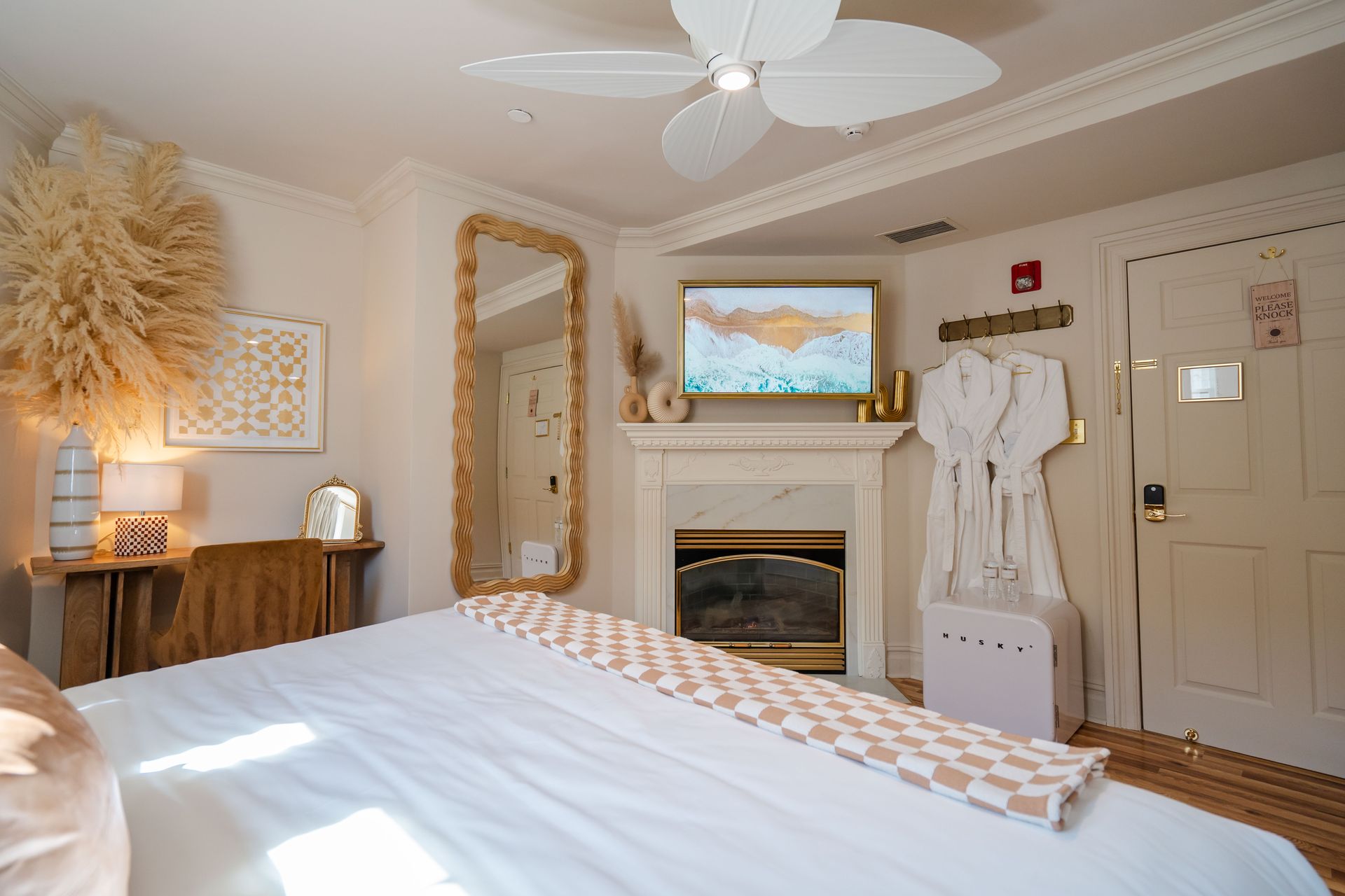 Cozy hotel room with a bed, fireplace, mirror, desk, and decorative dried grasses; off-white and gold hues.