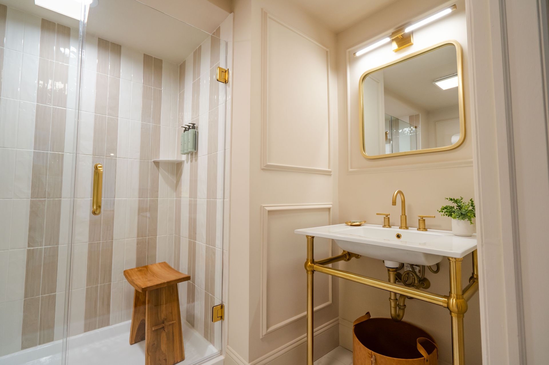 Elegant bathroom with gold fixtures, marble sink, and striped shower tiles; a wooden stool sits in the shower.
