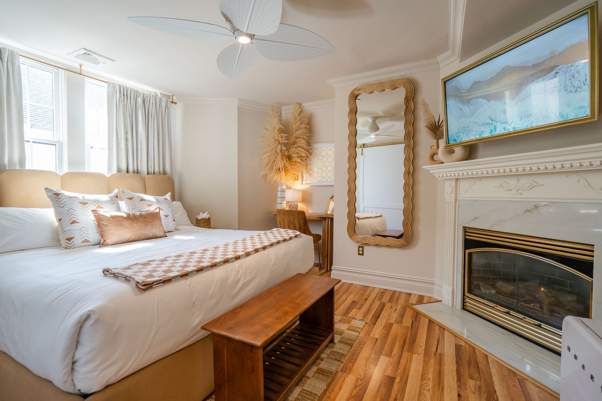 Cozy bedroom with a king-sized bed, fireplace, large mirror, and ceiling fan. Light wood floors and soft neutral tones.