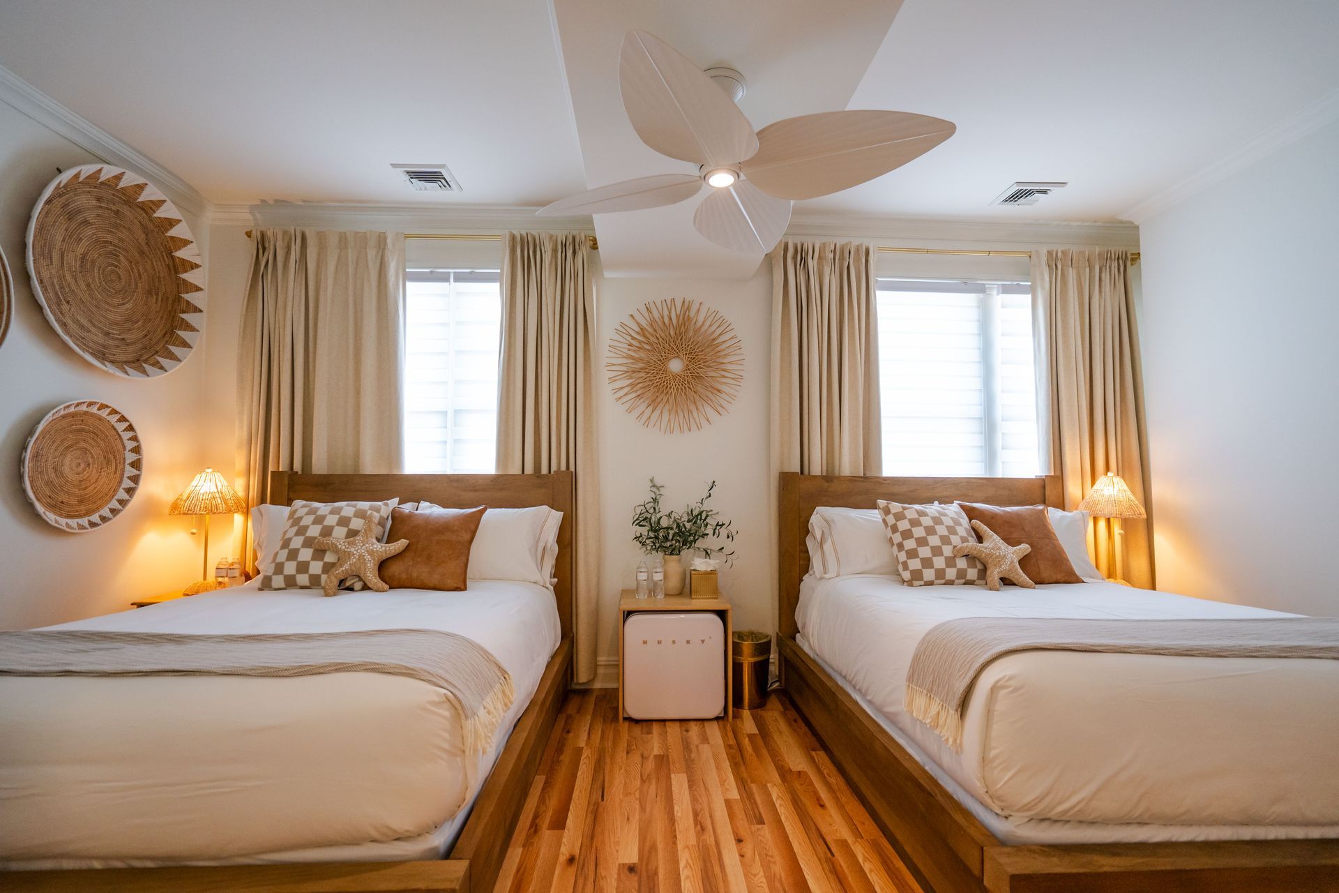 Two beds in a bright room with beige curtains and decorative woven wall art.
