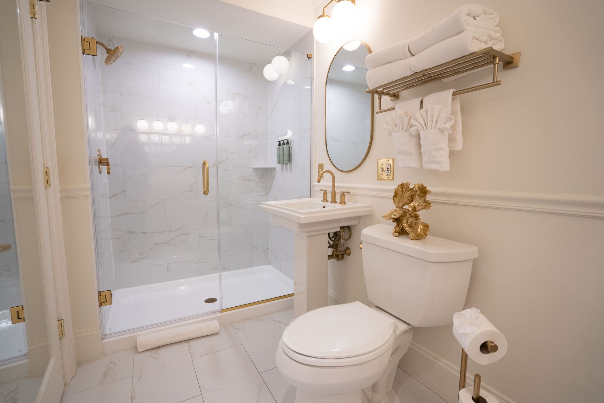 Elegant white bathroom with marble shower, gold fixtures, and a pedestal sink.