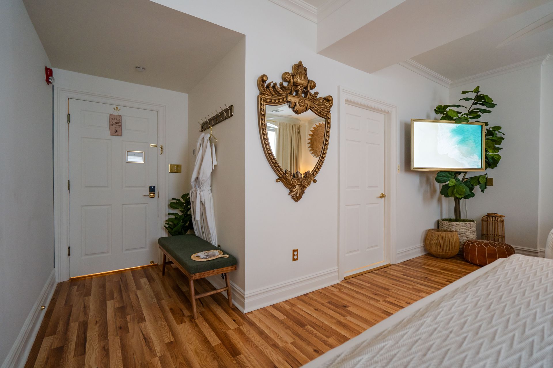 Hotel room entrance with white walls, wooden floors, ornate gold mirror, tv, bench, and potted plant.