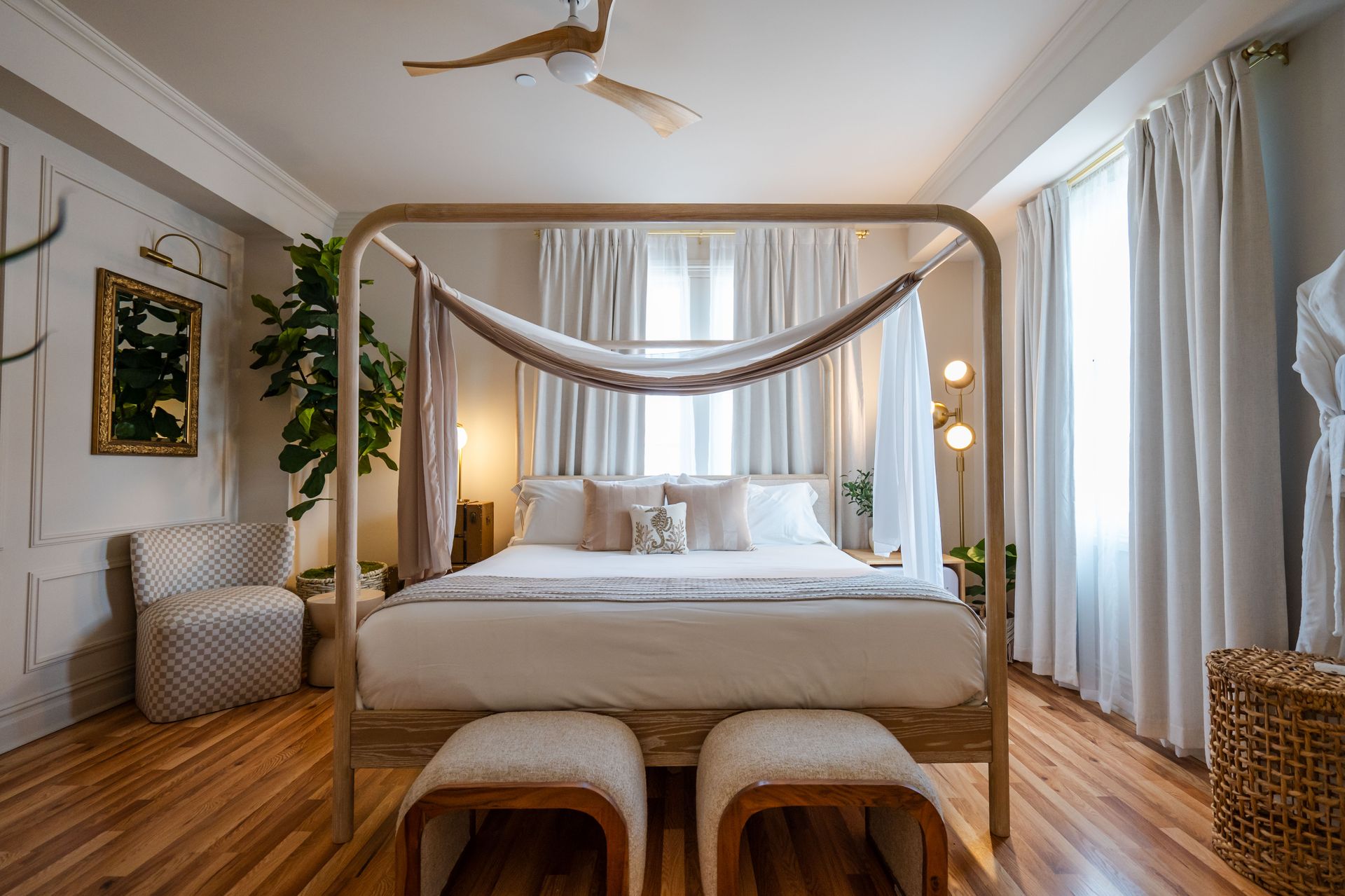 Bedroom with canopy bed, white bedding, wooden floors, sheer curtains, and two ottomans.