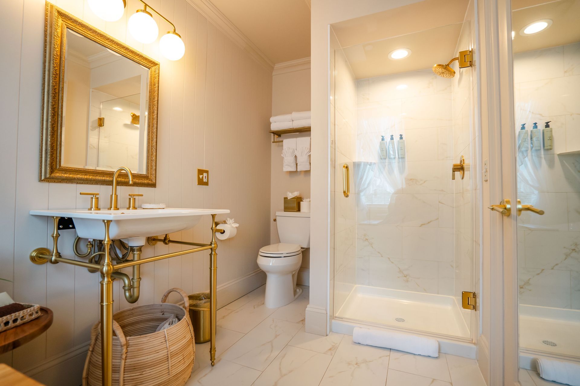 Bathroom with white walls, marble shower, and gold fixtures. Vanity with a gold frame mirror and sink.