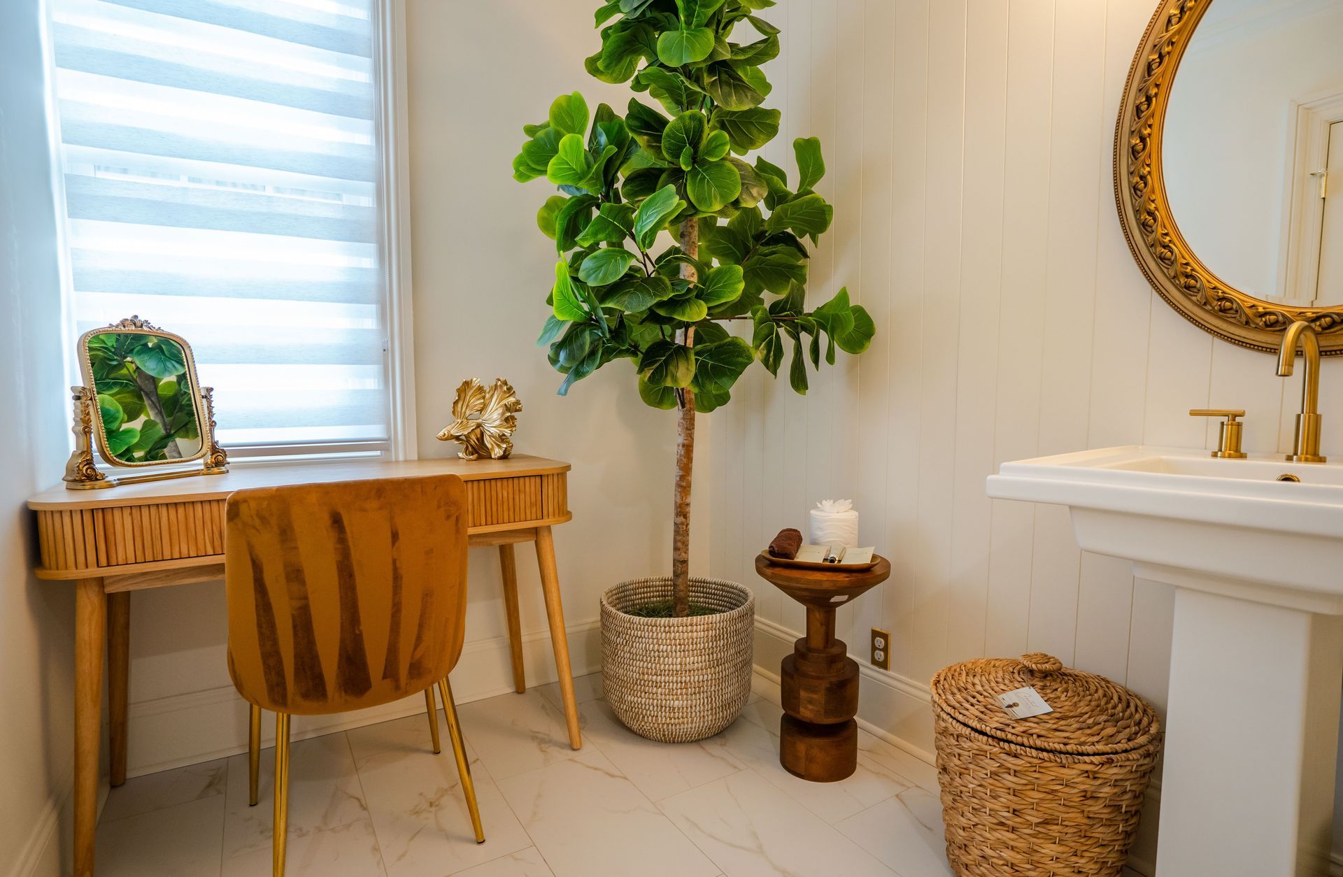 A bright bathroom with a desk, gold mirror, and large potted plant.