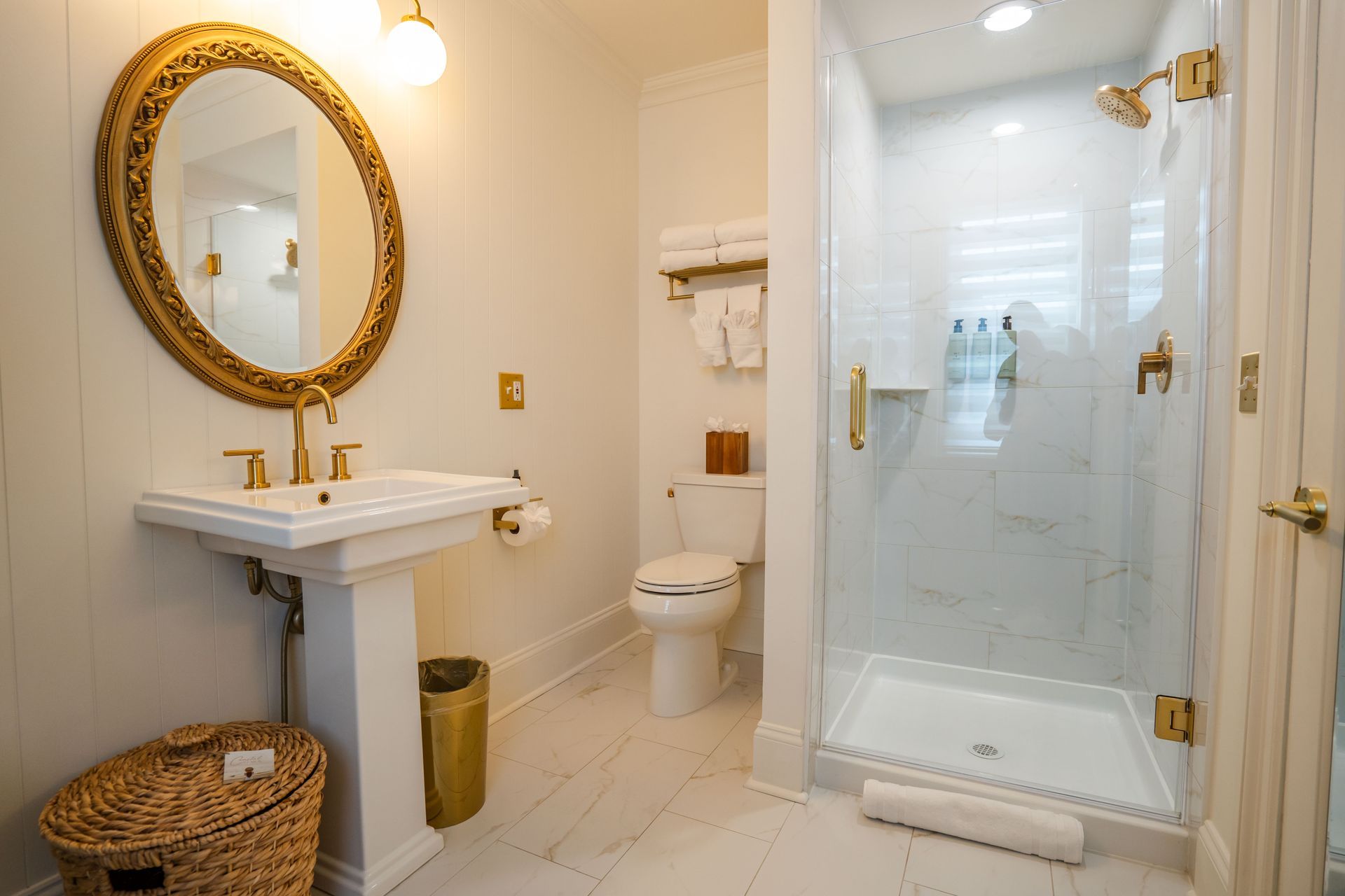 Bathroom with gold accents: pedestal sink, oval mirror, glass shower, white tile, and a wicker basket.