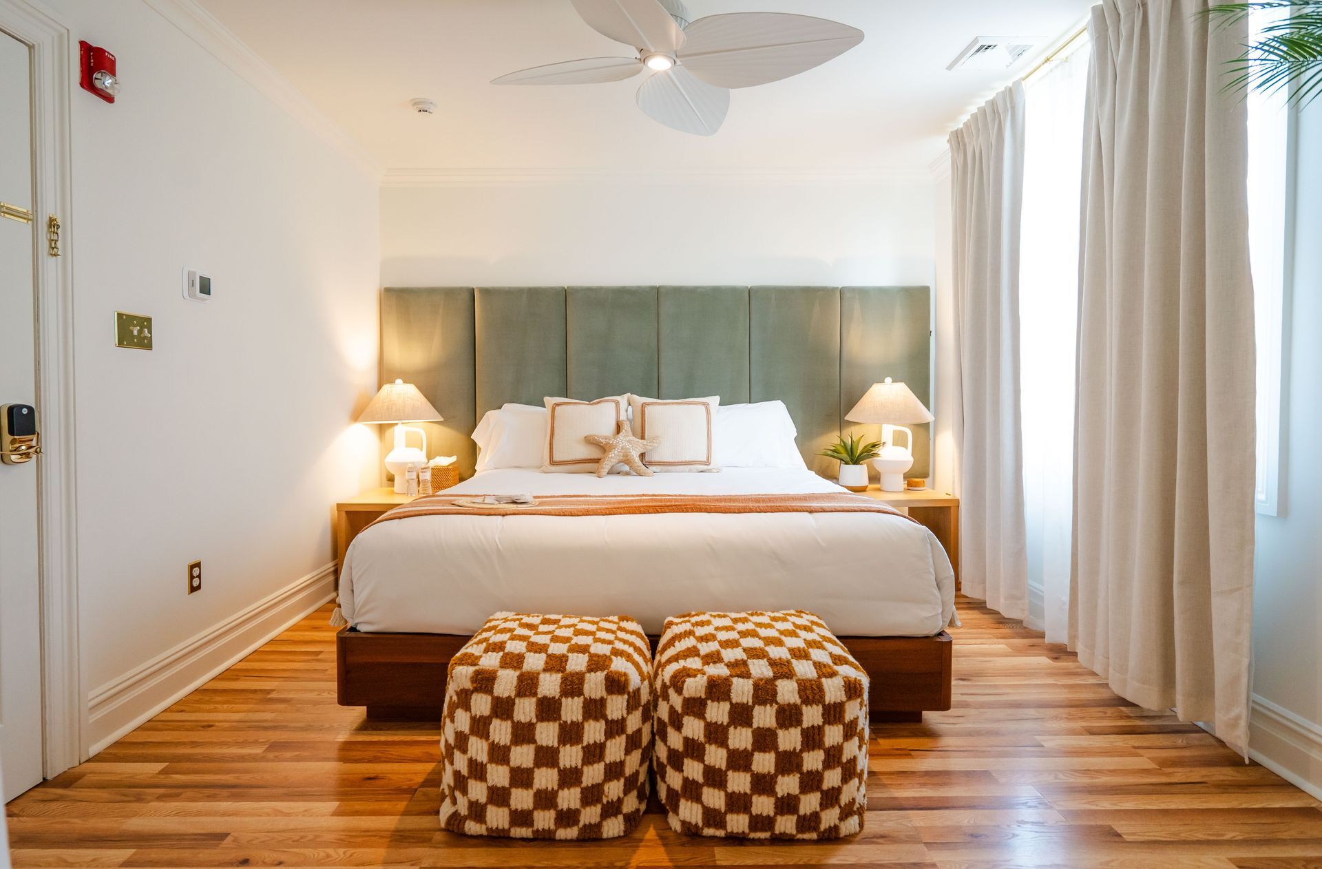 Bedroom with bed, checkered ottomans, and a sage green headboard. Wooden floor and white curtains.