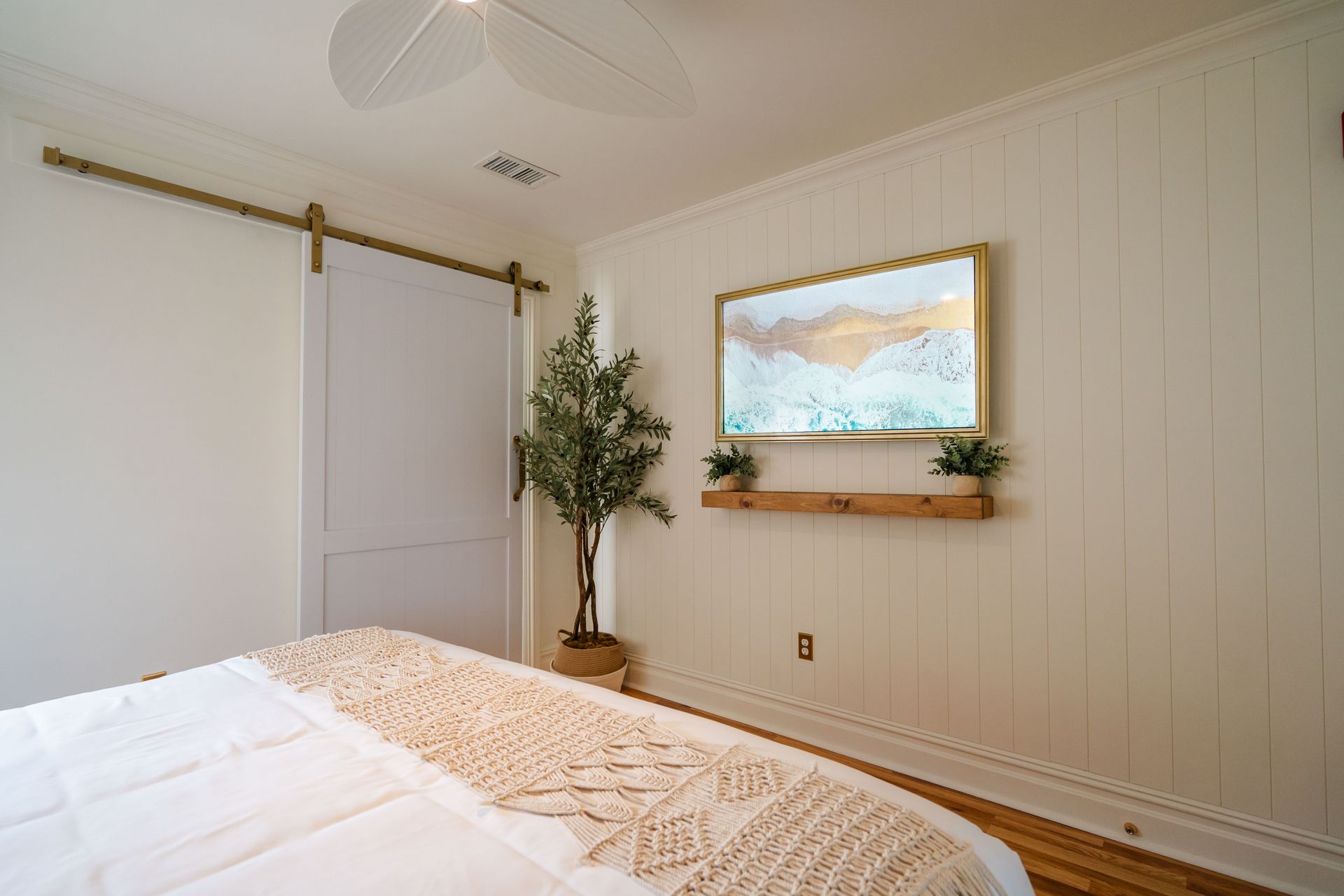 Bedroom with white walls, sliding door, art, plants, bed with cream-colored throw, and wood floors.