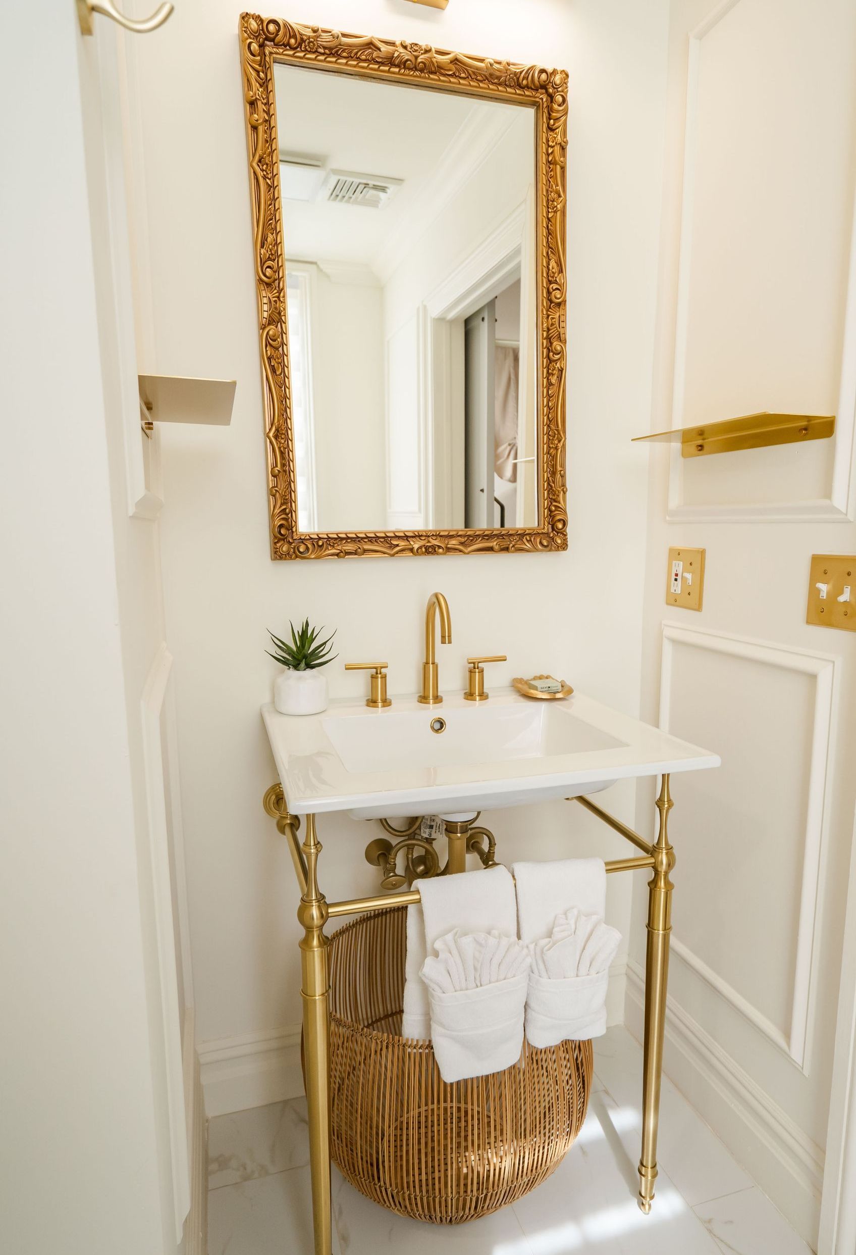 Elegant bathroom with gold accents: ornate mirror, faucet, and stand; white sink, walls, and towels, with a wicker basket.