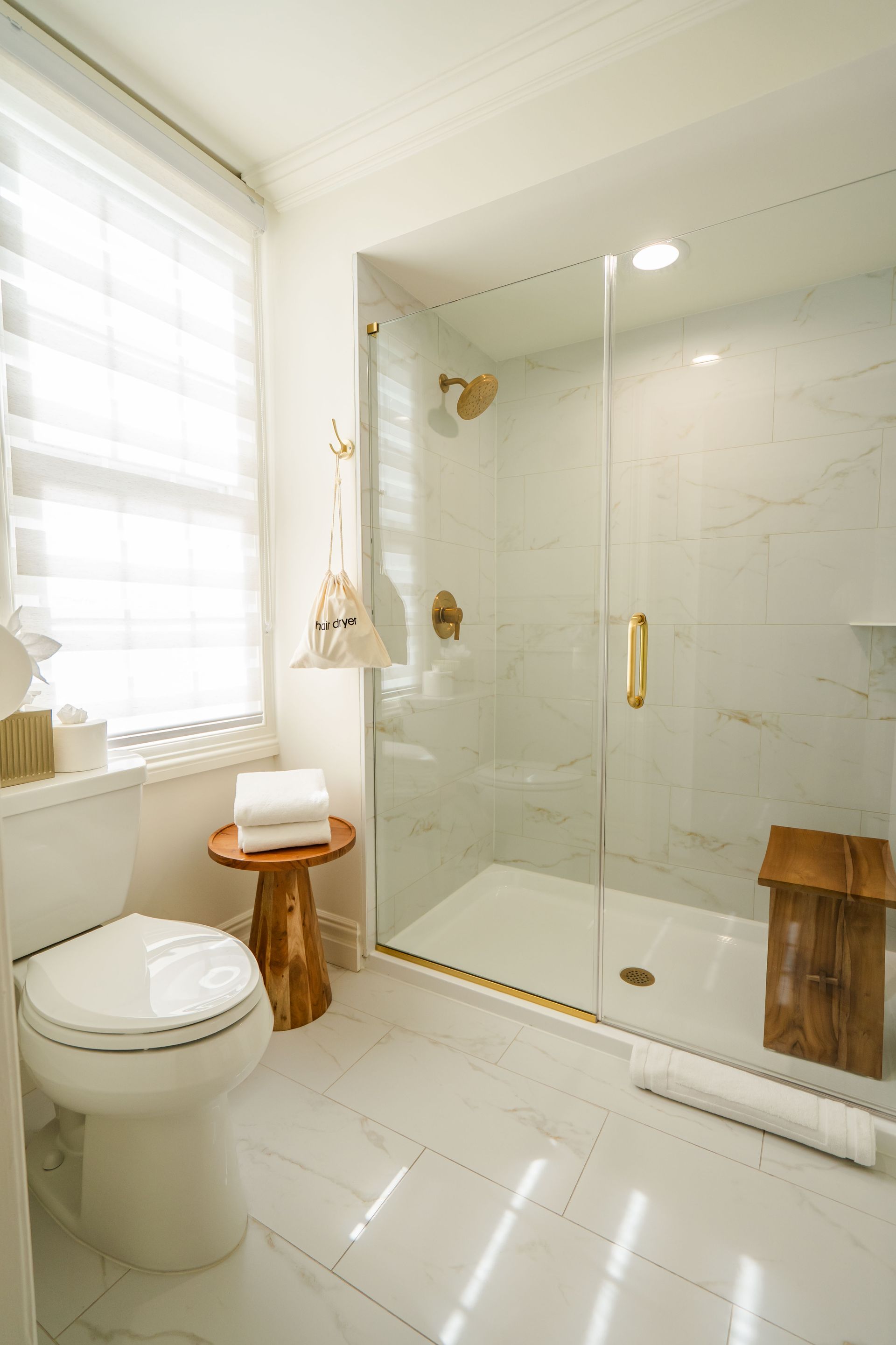 White bathroom with glass shower, gold fixtures, wood stool, and toilet.
